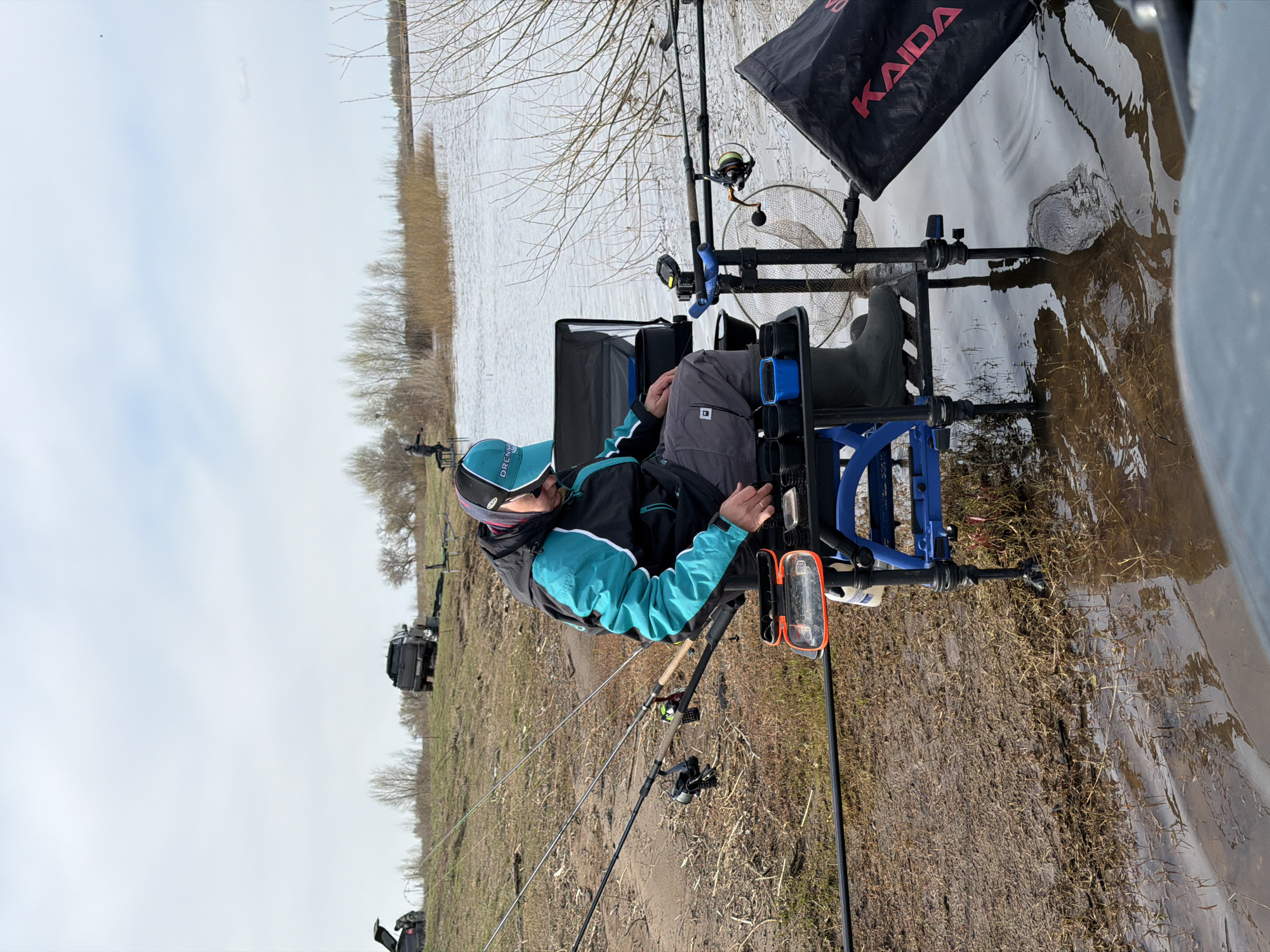 A person sits in a fishing chair by the water, potentially near Nikolo-Berezovka, Russia, given the context. The chair is equipped with fishing rod holders and various containers. Fishing rods are set up, extending towards the water. A black bag labeled "KAIDA" is also visible. The surrounding environment appears to be a muddy shoreline or riverbank.