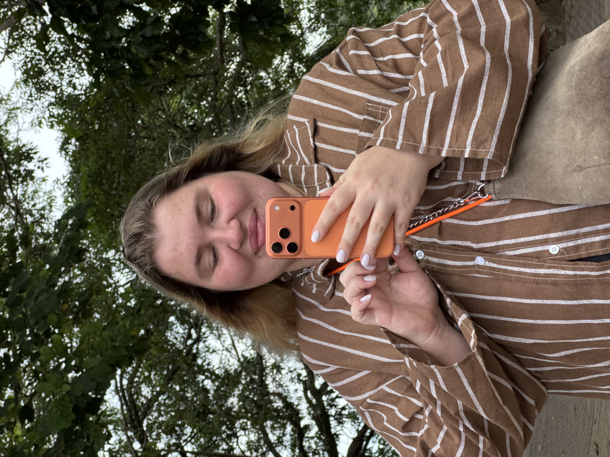 A woman with light brown hair, wearing a brown and white striped button-up shirt, stands outdoors and holds an orange smartphone in her hands, looking at the screen. Her eyes are closed, and her lips are slightly parted in a gentle smile. Her fingernails are painted white. Behind her, lush green foliage and tree branches are visible against a slightly overcast sky. A beige handbag with an orange strap hangs from her shoulder. The location is Karon, Thailand.