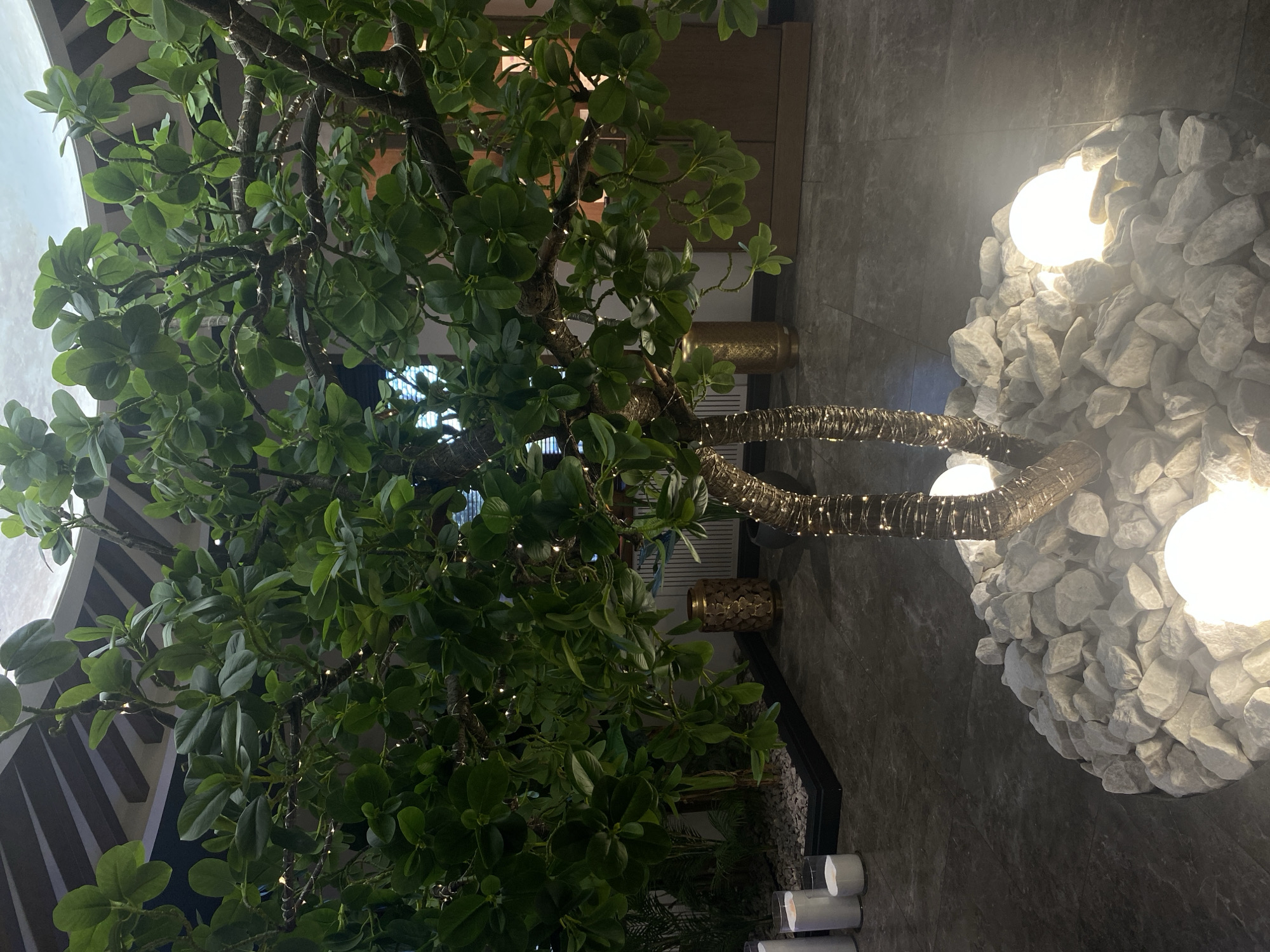 This image captures an indoor decorative setting, rotated 90 degrees clockwise from its natural upright position. The central subject is a large, artificial tree with abundant, dense green leaves. Numerous small, twinkling fairy lights are intricately wrapped around its branches, casting a soft glow. The tree's main trunks are also adorned with a textured, possibly metallic or golden, wrapping.
At the base of the tree, a substantial arrangement of white, irregularly shaped rocks is visible. Embedded within these rocks are two prominent spherical lights, emitting a bright, warm white illumination. The background features dark grey, possibly tiled, walls or flooring, providing a muted contrast to the tree's greenery and lights. In the mid-ground, glimpses of decorative golden or brass planters can be seen. Further back, architectural elements with dark, geometric lines are partially visible. Towards the bottom of the frame (which would be the left side if rotated correctly), several white cylindrical objects, resembling large candles or decorative accents, are present.
No people are visible in the scene. The overall ambiance is tranquil and sophisticated, typical of a modern interior space such as a hotel lobby, restaurant, or commercial building designed for aesthetic appeal. The extensive use of artificial lighting, from the fairy lights to the glowing spheres, suggests an evening or nighttime setting, or an interior environment where artificial illumination defines the mood. No visible text is present. The location is Votkinsk, Russia.