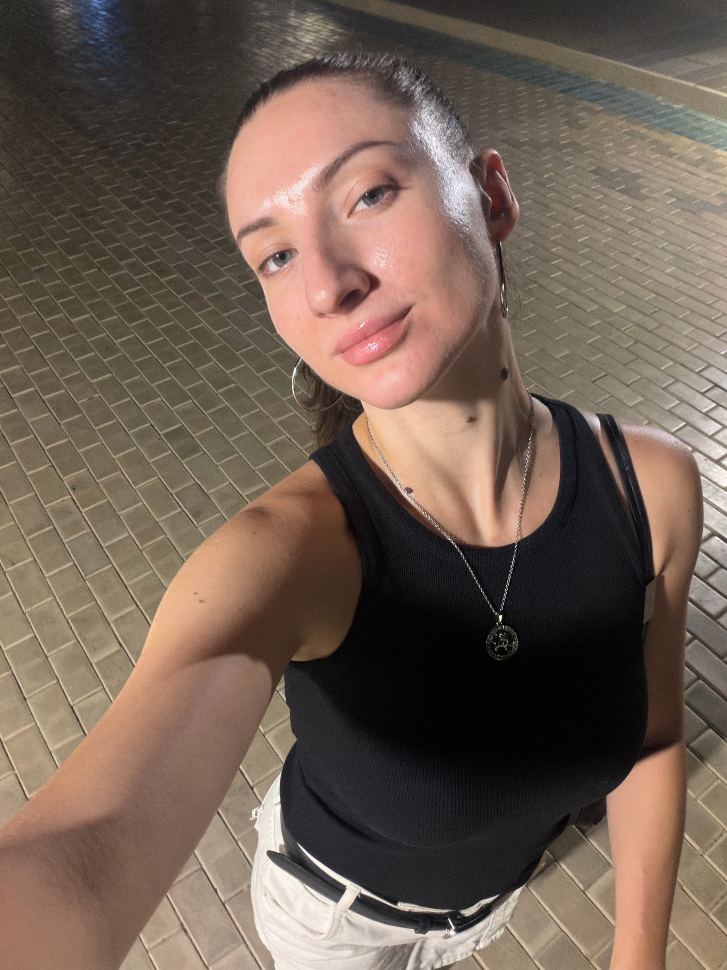 A medium-full shot taken at night in Sharm El Sheikh, Egypt, shows a young woman with her face partially illuminated by overhead lighting. She is looking upwards towards the camera with her head tilted slightly. Her hair is pulled back into a ponytail. She is wearing a black sleeveless tank top and white pants. A silver necklace with a circular pendant is visible around her neck. The ground is paved with light-colored rectangular tiles, and there are visible lines of light reflecting on the wet surface of the pavement. The background is out of focus, suggesting a street or outdoor area.