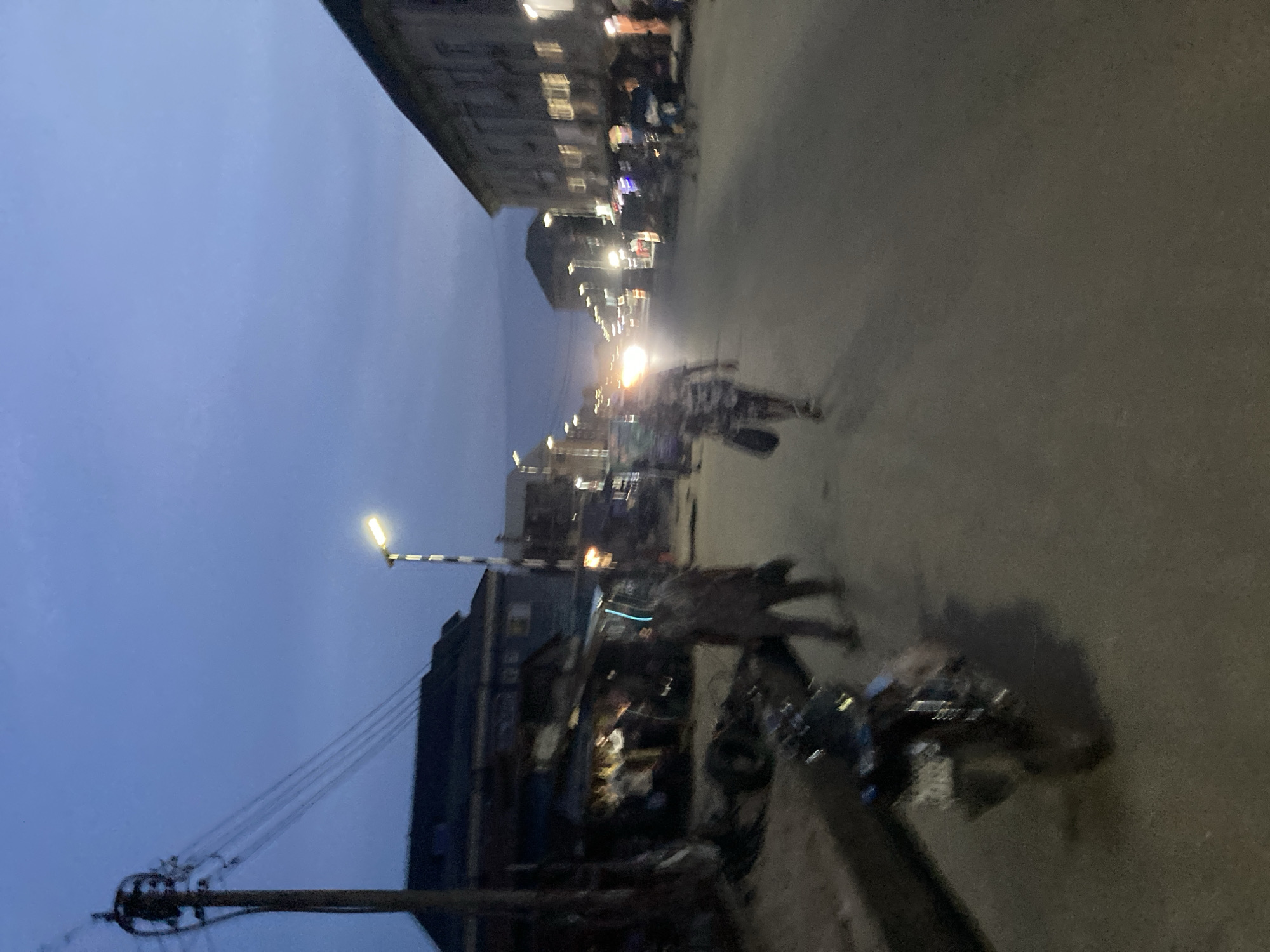 The image depicts a street scene at dusk or dawn in Aba, Nigeria. The sky is a deep blue. A street light illuminates the foreground. Buildings line both sides of the road, with the structures appearing more densely packed on the left. Several motorcycles are parked along the right side of the road. People are walking or riding bicycles on the street, visible as blurred figures. The road surface is paved. Power lines are visible extending from a utility pole. Distant headlights indicate vehicular traffic further down the road. The overall scene suggests a bustling urban environment in the early morning or late evening.