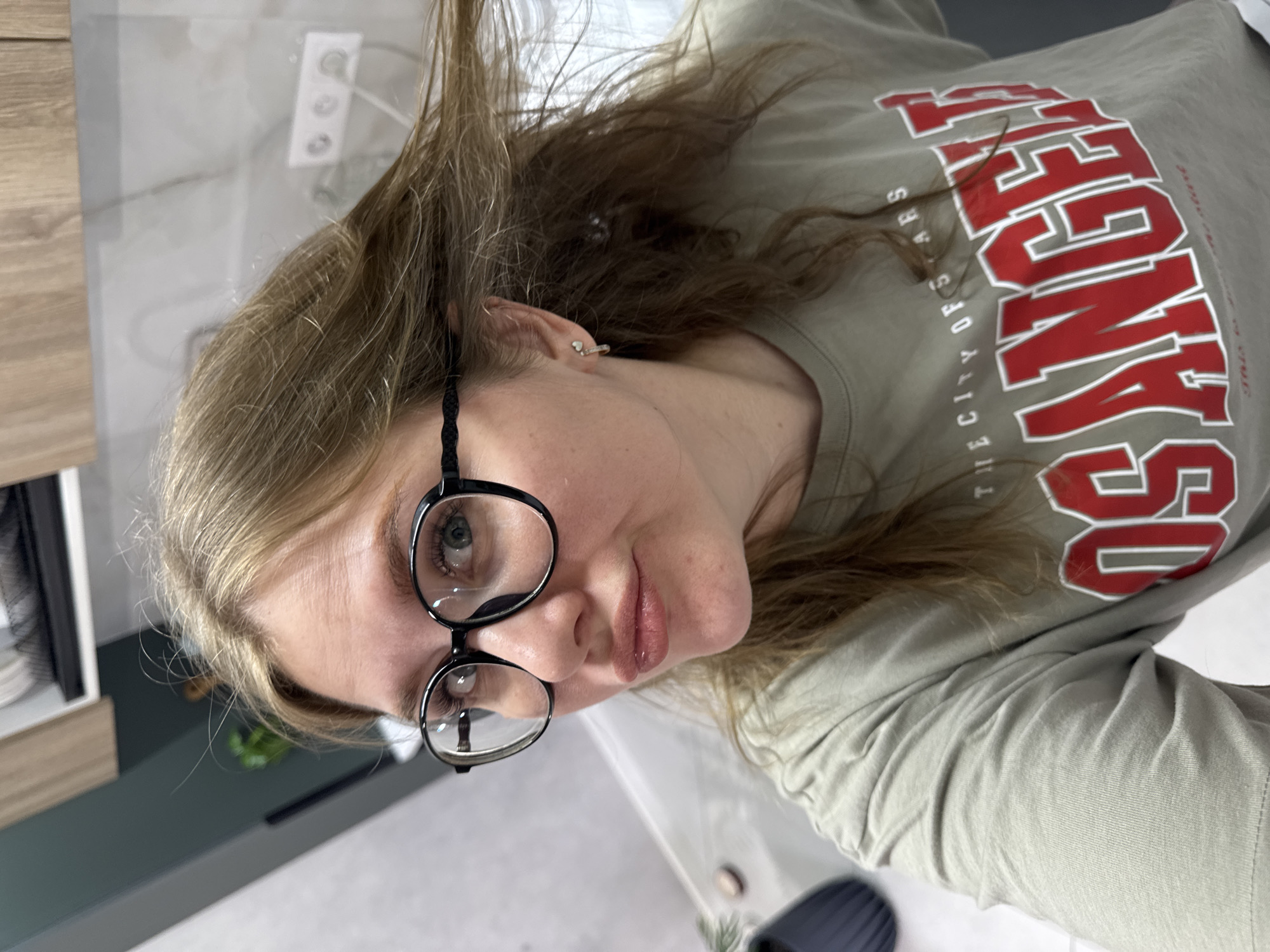 A woman with light brown hair and blue eyes, wearing black-framed glasses and a light olive green t-shirt with red and white lettering that reads "LOS ANGELES THE CITY OF STARS," is positioned indoors. She is looking directly at the camera with a slight smile. The background shows a kitchen or bathroom setting with a wooden cabinet to the left and a white tiled wall with an electrical outlet in the upper left. A small plant is visible on a dark green surface to the left. The location appears to be indoors, but the city of Gornyi (Dvurechenskoe S/P), Russia cannot be determined from the image.