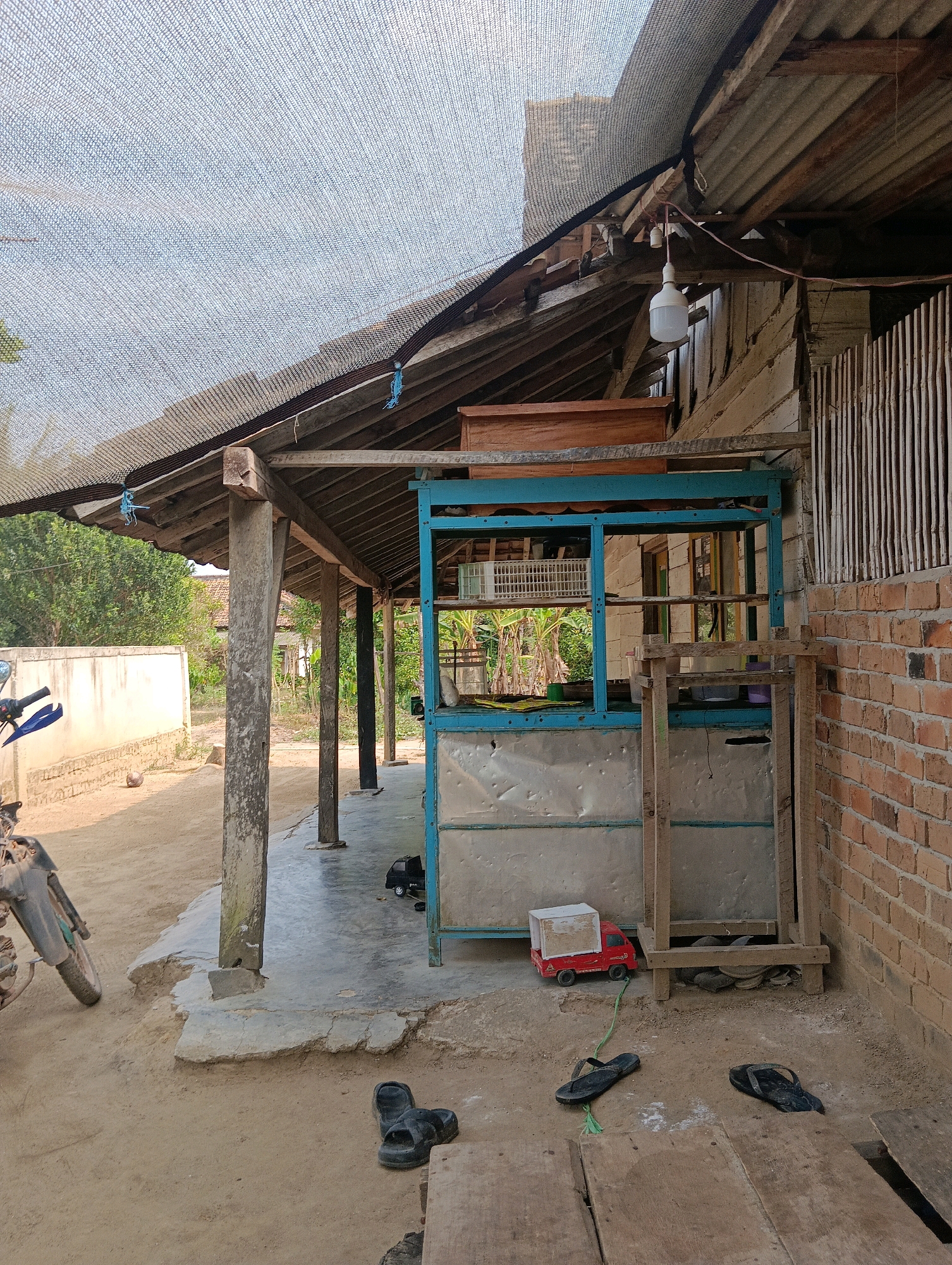 The image depicts an outdoor scene at a small shop or stall in Binakarsa, Indonesia. A simple, open-air structure with a corrugated metal roof supported by wooden posts and a tarp overhead provides shade. In the foreground, a motorcycle is parked on a dusty ground. To the right, a brick wall forms the side of an adjacent building. In the center of the scene is a blue metal stall with various items, including a white basket and what appears to be a toy red truck. Several pairs of black flip-flops are scattered on the ground around the stall. The environment suggests a casual, everyday setting, likely in a residential or market area.