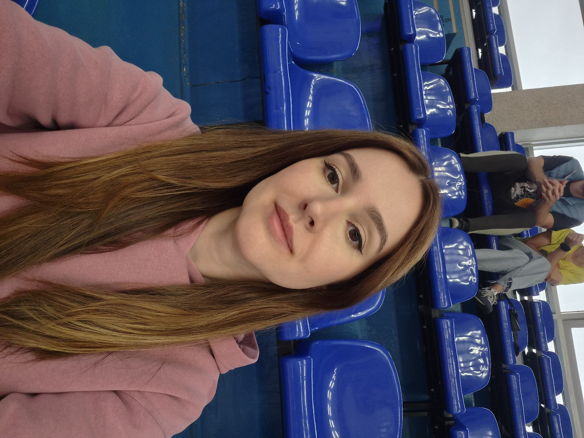 A young woman with long, light brown hair is taking a selfie while sitting in an empty row of blue stadium seats. She is wearing a pink hooded sweatshirt. The background consists of several rows of blue seats and a glimpse of people sitting in the rows behind her. The lighting suggests an indoor or overcast setting. The location is not identifiable from the image.
