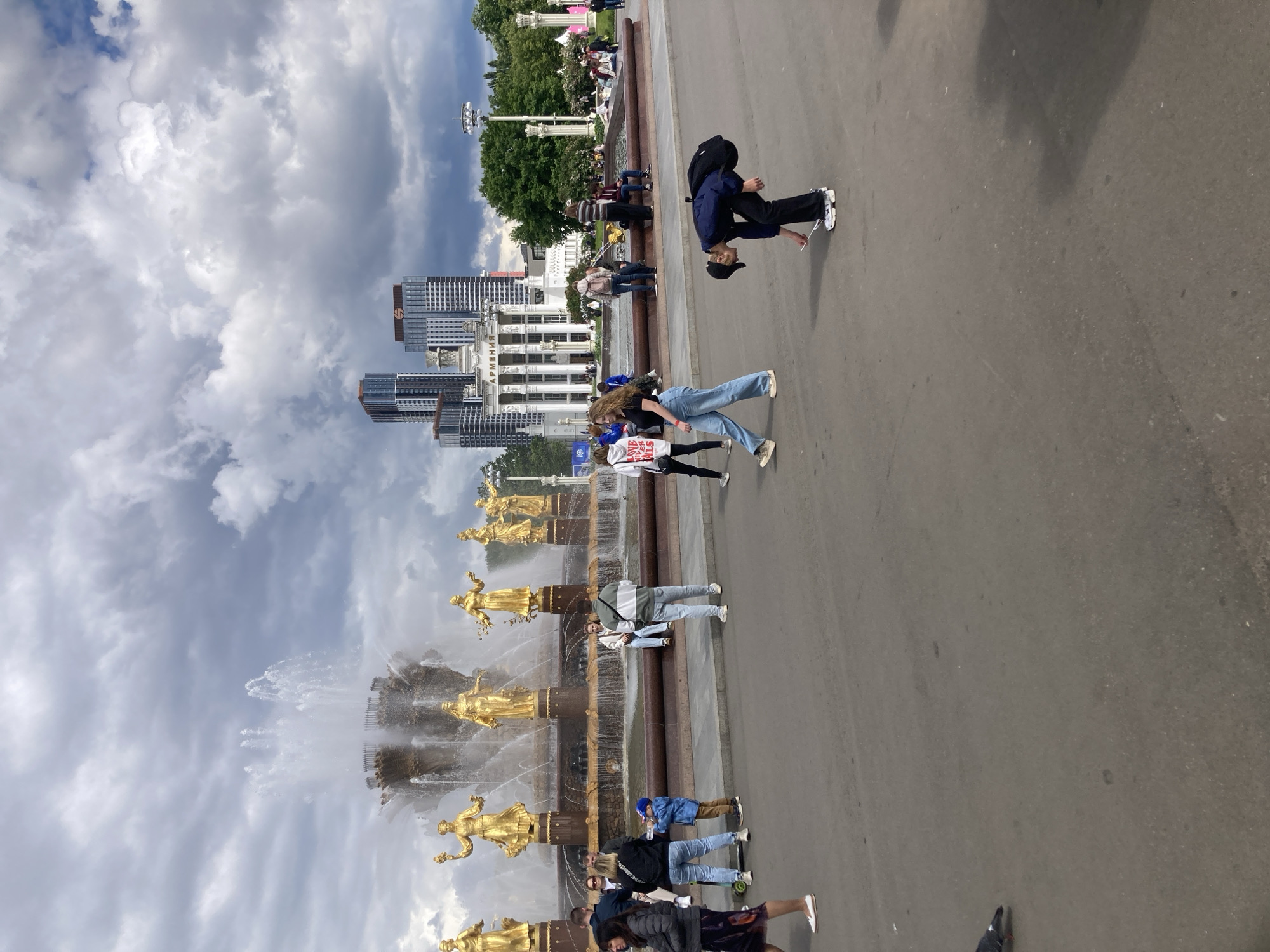 The image depicts a daytime scene in Moscow, Russia, specifically at the Exhibition of Achievements of National Economy (VDNKh). The focal point is the Friendship of Nations Fountain, characterized by its golden statues and cascading water. Numerous people are present, some walking along the pedestrian area adjacent to the fountain, and others interacting with the fountain itself. The architecture of the exhibition grounds is visible in the background, including buildings with distinct Soviet-era design elements. The sky is overcast with clouds.