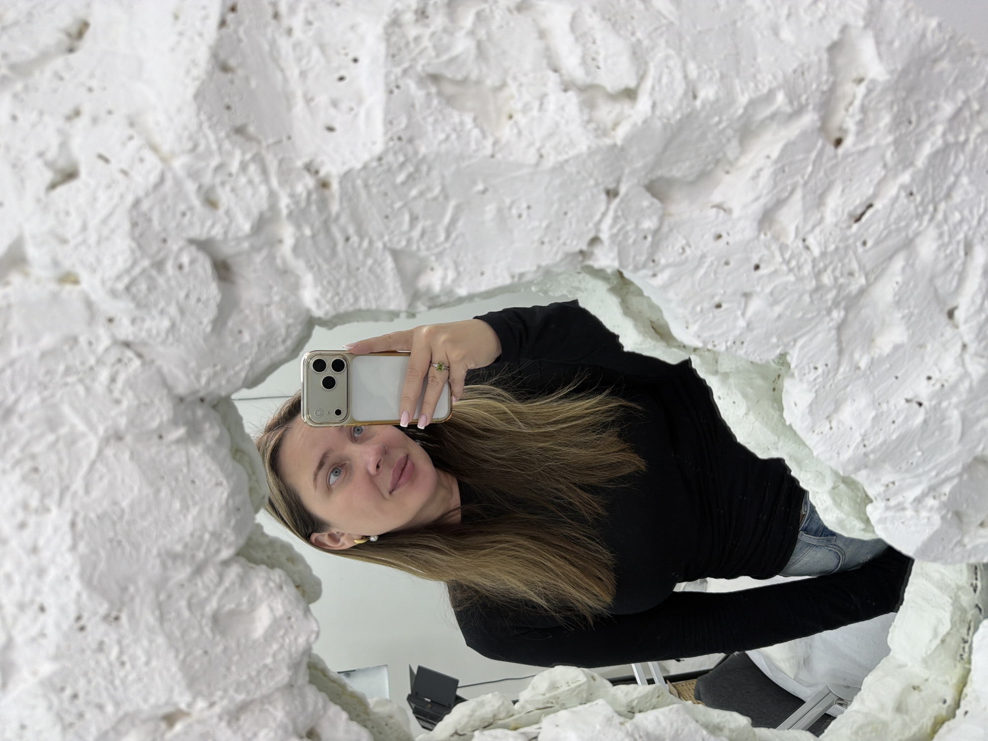 A woman takes a selfie in a mirror surrounded by white textured material. She wears a black long-sleeved shirt and jeans, holding a gold iPhone with a white back. Her hair is light brown, and she has on a ring. The mirror appears to be surrounded by decorative or sculptural elements with a rough, textured surface. The location is unknown.