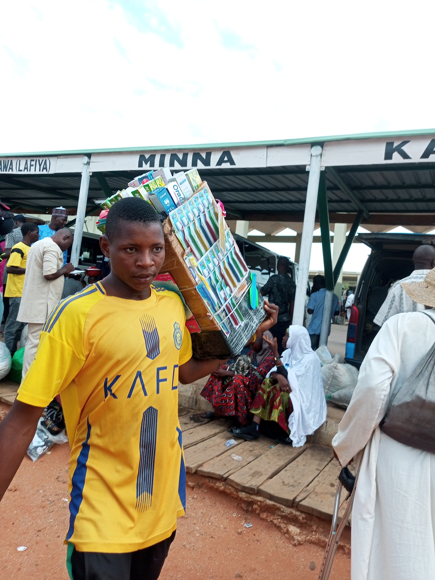 A young man is captured in a medium shot, carrying a large cardboard box filled with toothbrushes on his left shoulder. He is wearing a bright yellow t-shirt with blue stripes and a blue number '1' on the back, along with black shorts. The toothbrushes in the box are packaged in various colors, and some boxes have text such as "Oral-B" and "Crest."
Behind him, a covered bus station or market stall provides shelter. The text "MINNA KA" is visible on the side of the structure, suggesting a location or route. Several other individuals are present in the background, some sitting on wooden planks and others standing near vehicles. One person in the foreground on the right is holding a crutch, wearing a white robe and a straw hat. The ground is a mix of sand and dirt.
The scene appears to be outdoors during the day, with a bright, overcast sky. The overall atmosphere suggests a busy market or transport hub, possibly in Gombe, Nigeria, given the location context. The young man's expression is neutral, and he seems focused on carrying his load.