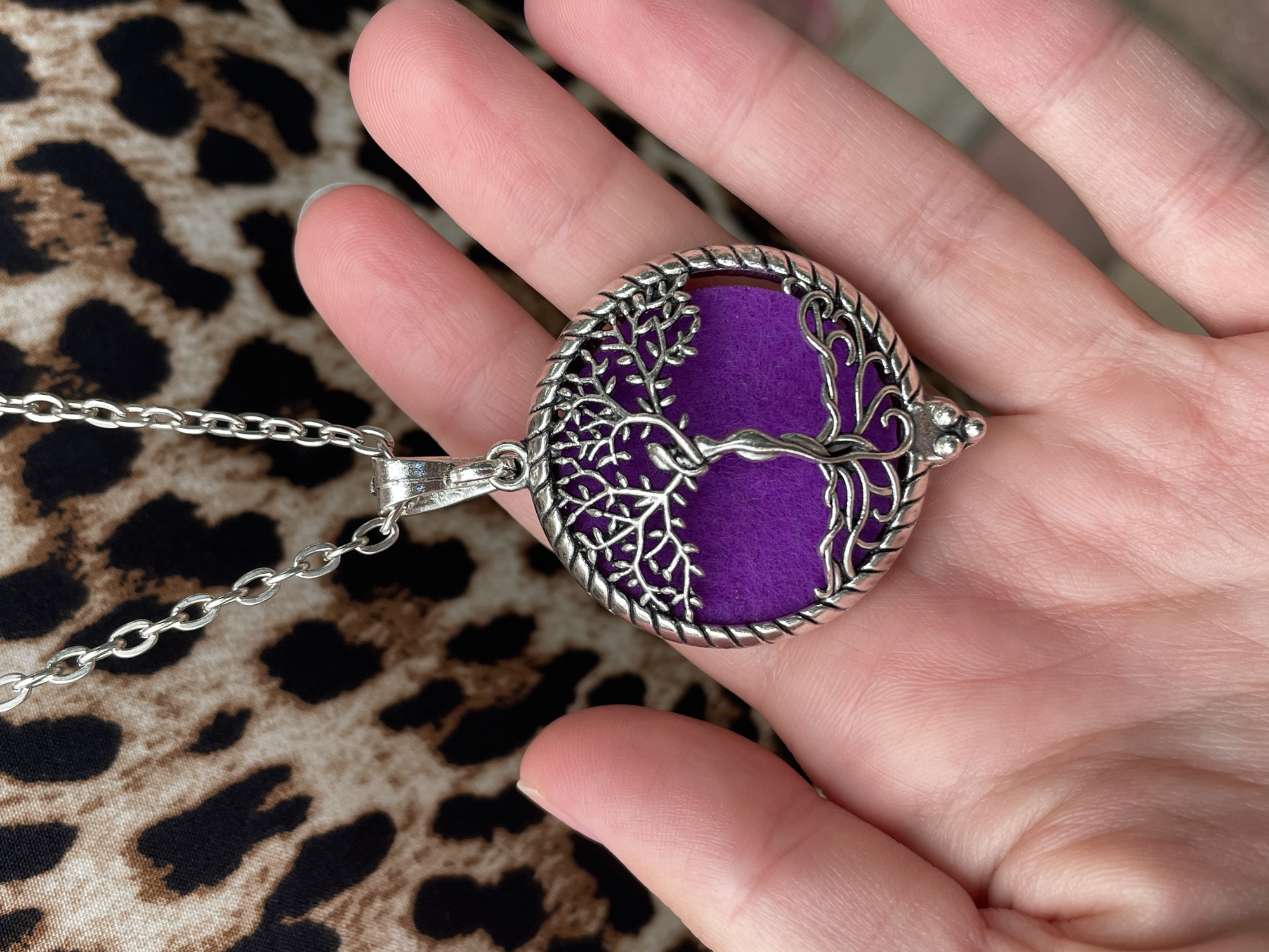 A hand is holding a silver pendant with a tree of life design and a purple circular insert, in front of a leopard-print fabric. The pendant is circular with a braided silver border. The tree of life is depicted in silver filigree in the center, with a female figure intertwined with the trunk. A silver chain is attached to the top of the pendant. The background is a leopard-print pattern in shades of brown and black. The image is well-lit, with the focus on the pendant and the hand.