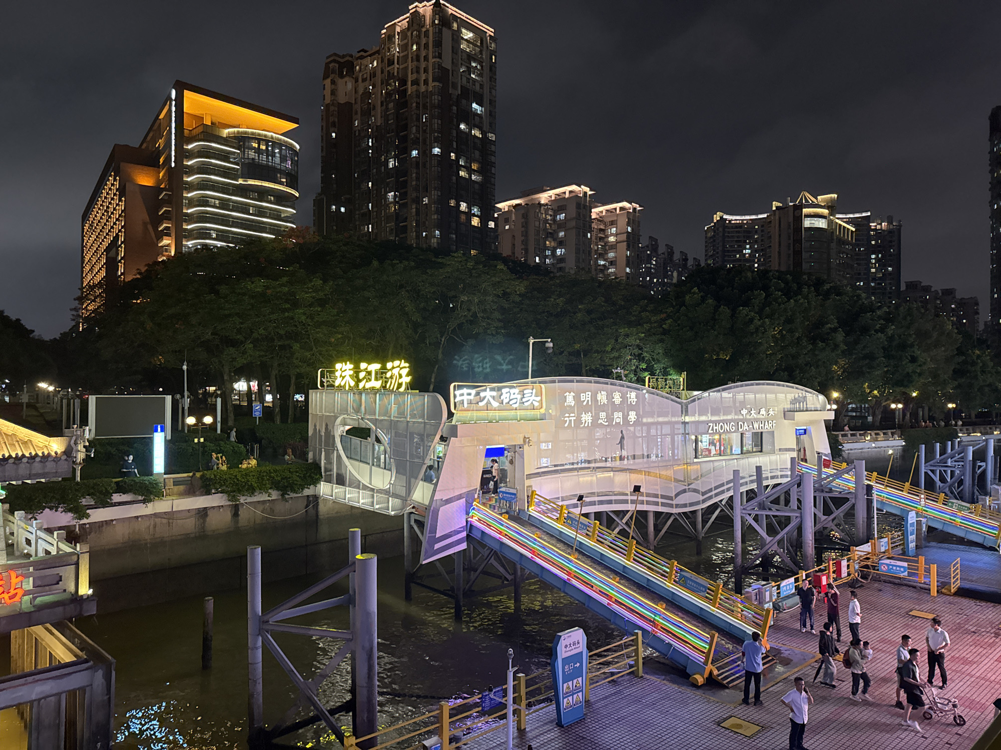 The image depicts a night scene in Haizhu District, China, featuring the Zhong Da Wharf, illuminated by colorful lights. The wharf is a modern structure with ramps leading to the water. A group of people are gathered on the dock, some walking, some standing. In the background, several tall buildings are visible, also lit up at night. Trees line the waterfront, and the dark water reflects the ambient light. Signage in Chinese and English identifies the location.