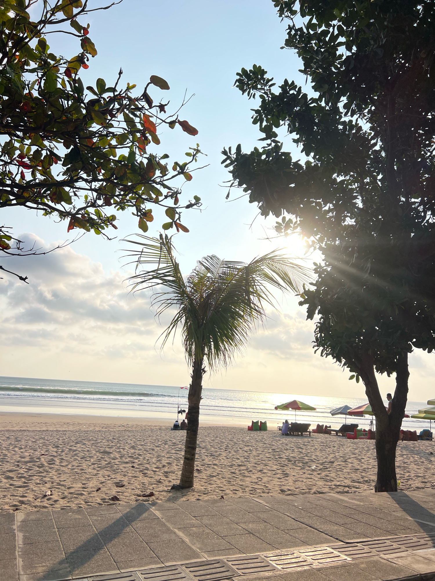 This image depicts a serene beach scene in Kuta, Indonesia, likely during the late afternoon or early morning. The foreground features a paved walkway with distinct textured tiles, casting long shadows across the sandy beach. Two large trees frame the scene on the left and right, their foliage partially obscuring the bright sky and sun. The left tree, with its green and reddish-brown leaves, has branches extending towards the sky. The right tree is denser, its dark leaves silhouetted against the bright light.
The sandy beach stretches towards the ocean, dotted with several colorful beach umbrellas and lounge chairs. Some figures are visible relaxing on the chairs or walking on the beach, suggesting a leisurely atmosphere. A lone palm tree stands in the mid-ground, its fronds swaying gently. The ocean is calm, with gentle waves rolling onto the shore, and the sun's rays create a shimmering effect on the water's surface. The sky is a mix of pale blue and soft white clouds, illuminated by the bright sun peeking through the tree branches.