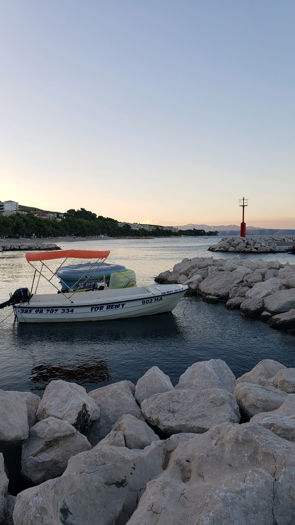 The image depicts a serene marina scene in Općina Tučepi, Croatia, at dusk. A white boat, labeled "FOR RENT," is moored in the foreground, its orange canopy casting a soft shade. The boat displays registration numbers "385 98 707 334" and "902 MA." To the right of the boat, a breakwater constructed of large, light-colored rocks extends into the water. At the end of the breakwater, a red navigational light stands as a beacon. In the background, a calm sea meets a pale, clear sky, with a distant shoreline dotted with buildings and lush greenery. The overall atmosphere is tranquil, suggesting the end of a day at the coast.
