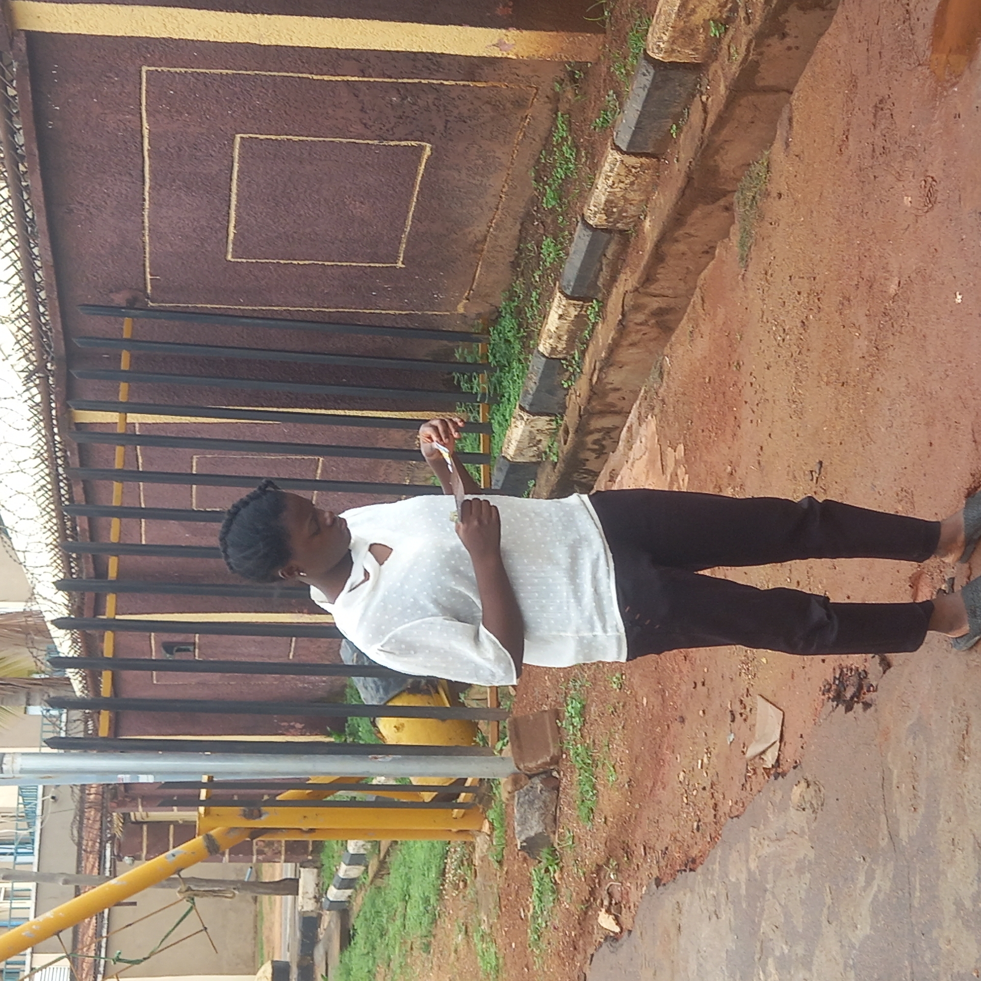 A young African woman is standing outside on a reddish dirt ground next to a concrete curb lined with black and white stone. She is wearing a white, polka-dot blouse and black pants. Behind her is a brown wall with a square design outlined in gold. There is a black iron fence with yellow accents in front of the wall. Based on the environment, this does not appear to be Barden Yana, Nigeria.