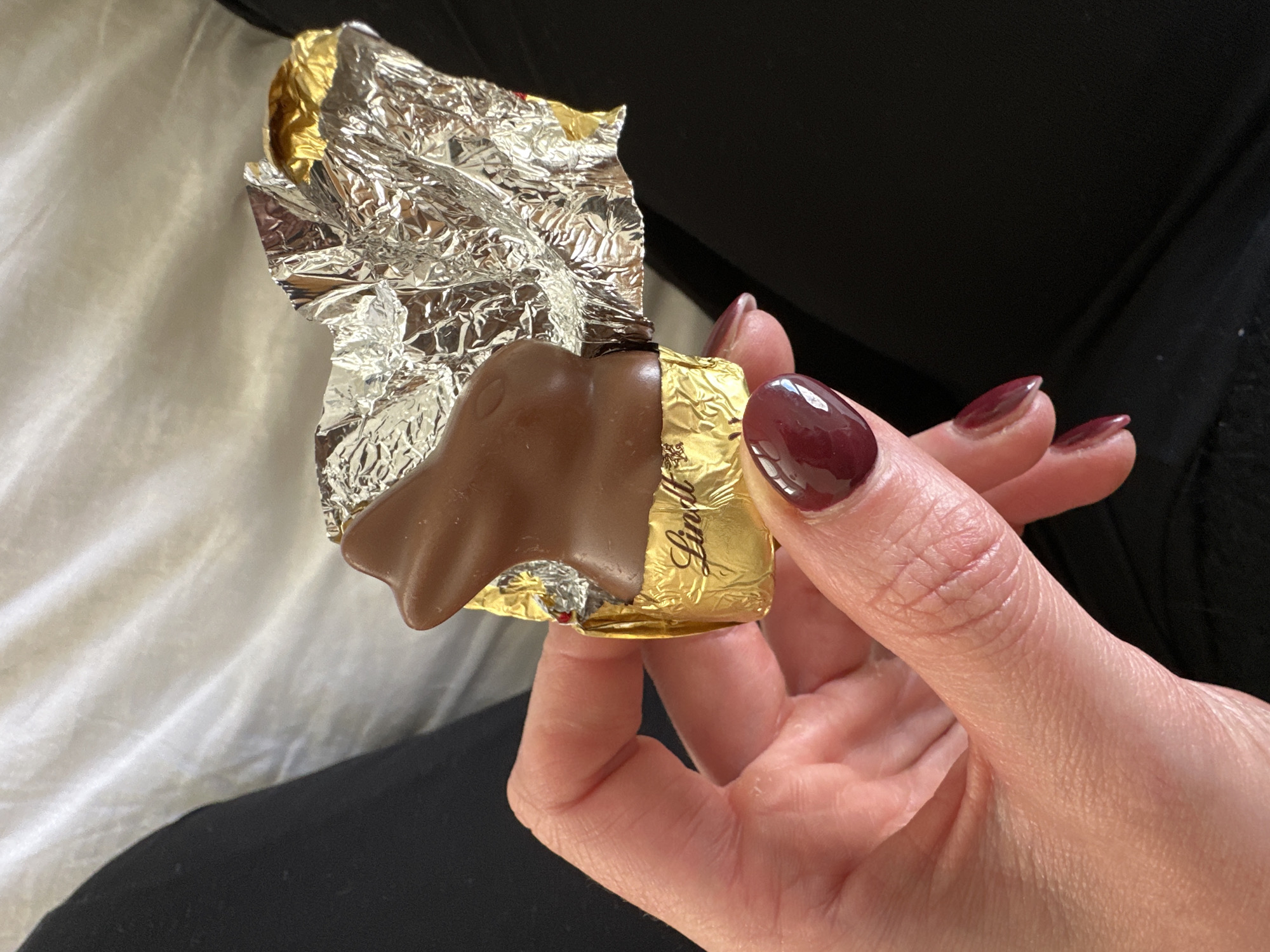 A hand with burgundy nail polish is holding a partly unwrapped Lindt chocolate bunny. The silver foil wrapper is partially open, revealing the brown chocolate underneath. The gold foil with the Lindt logo is still partially covering the chocolate. The background includes white bedding and a dark garment. The location is not discernible from the image, and there is no indication of the city of Wealden, United Kingdom.
