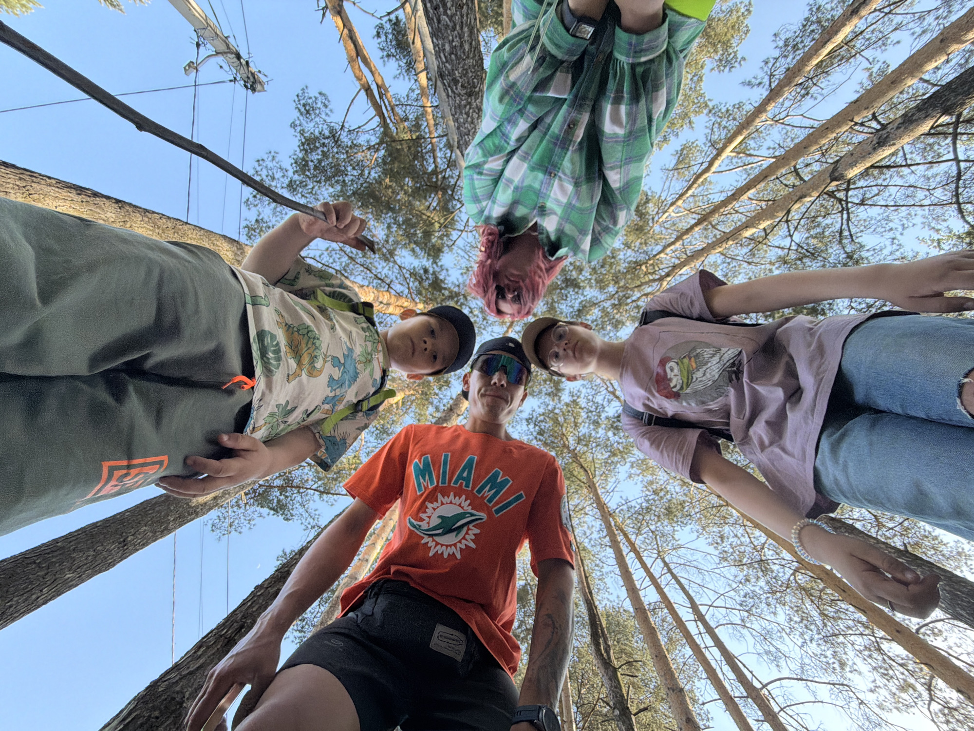 Five people are captured in a low-angle shot, looking down at the camera. The setting appears to be a forest, evidenced by the tall trees surrounding them, reaching up toward a blue sky.
From left to right: The first person wears a patterned shirt and green pants, holding a stick. The second person wears a dark baseball cap. The third person wears a Miami Dolphins t-shirt. The fourth person has pink hair. The fifth person wears a light purple shirt and light blue jeans.
The location of the photo is Chaikovsky, Russia.