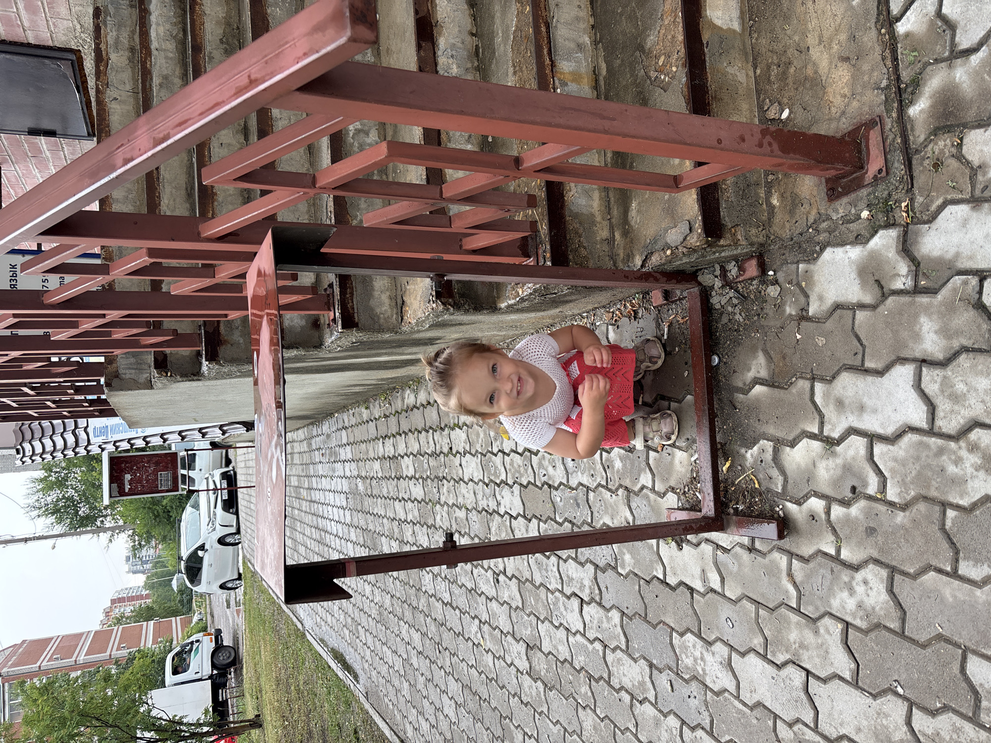 A young girl crouches within a metal frame on a wet, paved sidewalk. She is wearing a white knitted shirt and a red skirt. Her hair is pulled back in a ponytail. The frame appears to be part of a trash receptacle. To her right, a set of concrete stairs with a red metal railing ascends. In the background, a white truck and a white car are parked on the street, with apartment buildings visible behind them. The scene is located in the city of Blagoveshchensk, Russia. The overall lighting suggests an overcast day.