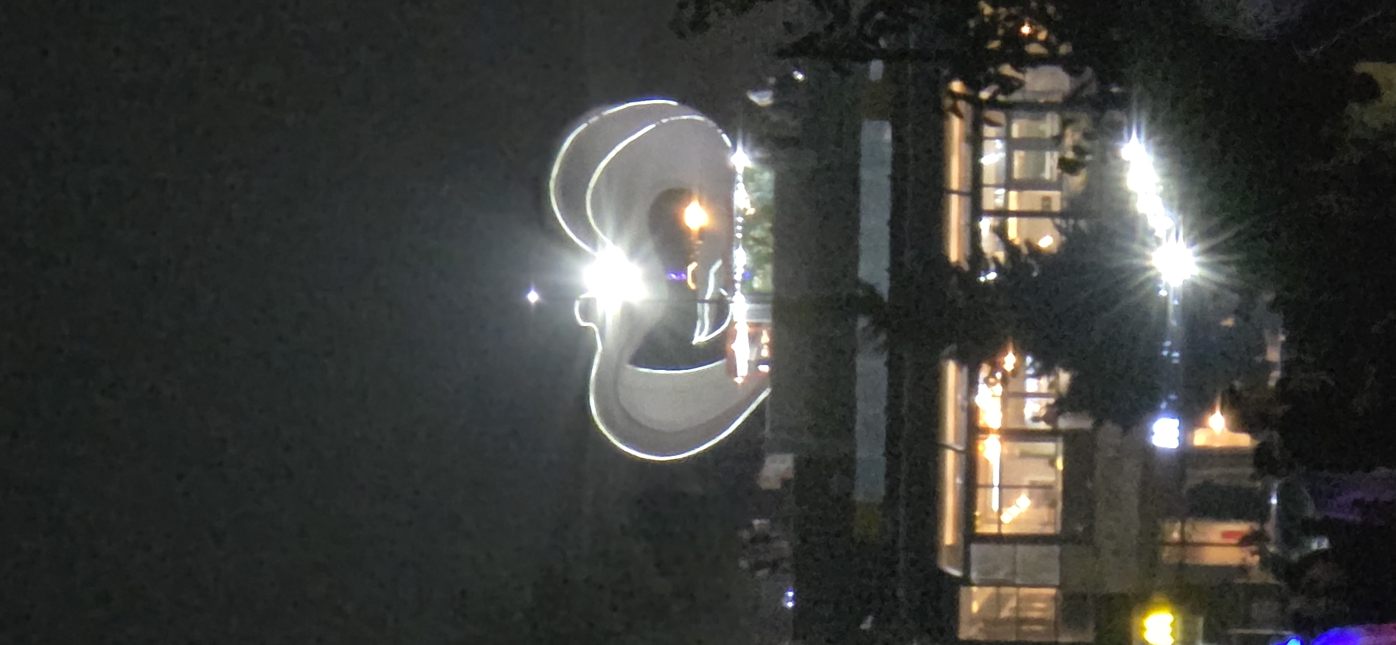 The image depicts a nighttime scene in Sumgayit, Azerbaijan, featuring a large, illuminated heart-shaped structure dominating the upper portion of the frame. Below this, a building with large windows glows with warm interior light, suggesting it is occupied or operational. In the foreground, silhouetted figures are visible amidst dark foliage, with bright streetlights and vehicle headlights creating a starburst effect. The overall ambiance is urban and festive, with the prominent heart structure possibly indicating a decorative or symbolic installation.