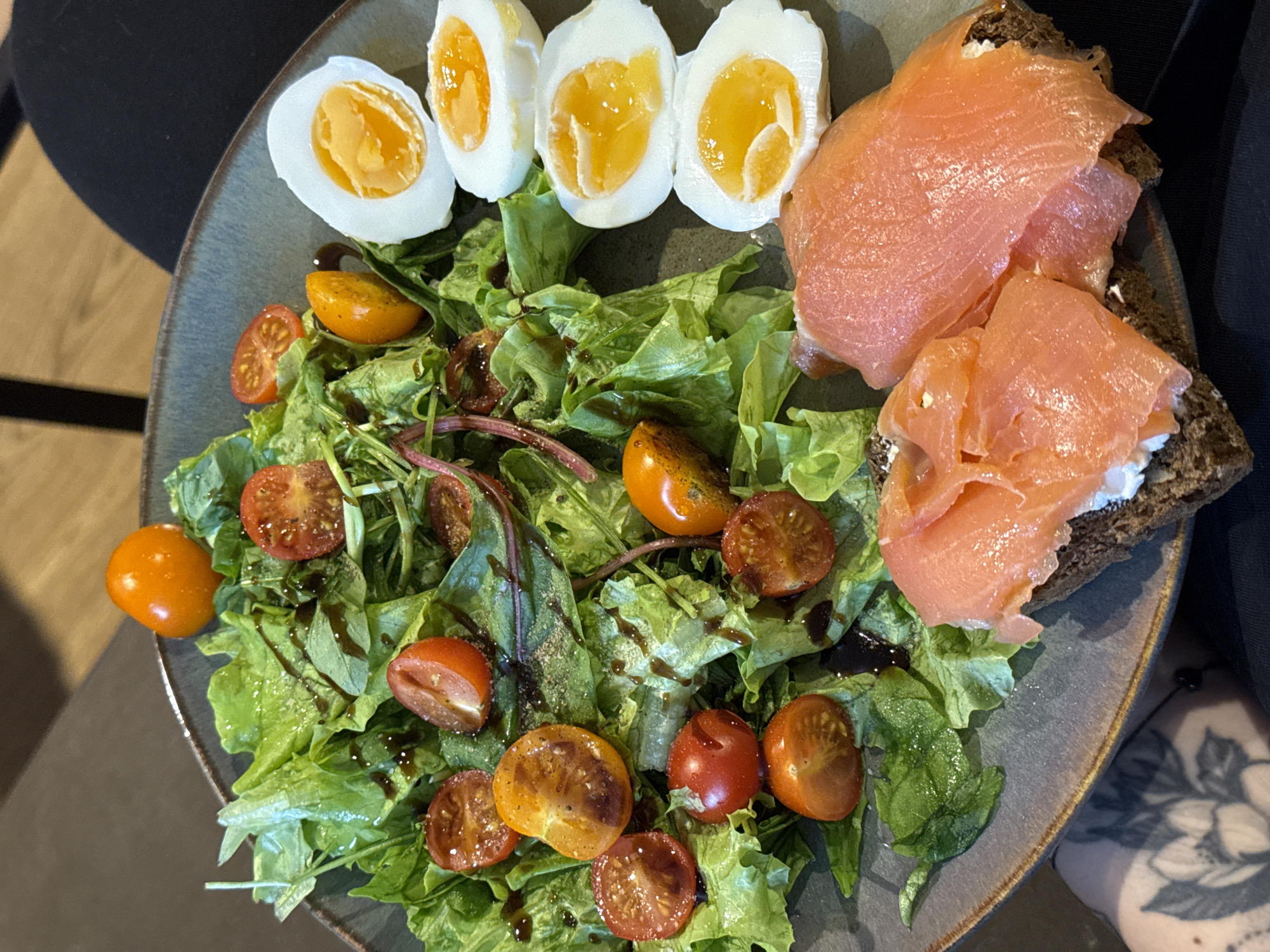 A top-down shot shows a plate of food containing a salad, eggs, and smoked salmon on bread. The salad consists of green lettuce, halved red and yellow cherry tomatoes, and is drizzled with a dark balsamic glaze. To the top left of the salad are three halved soft-boiled eggs with bright orange yolks. To the upper right of the salad is a piece of dark bread topped with cream cheese and several slices of smoked salmon. The plate is a muted blue-grey ceramic with a slight metallic rim. The setting is indoors and the plate is on a grey table. Parts of a person wearing a dark shirt are visible on the top left and right of the image, with a partial view of a tattoo on the wrist of the person on the lower right. There is no indication that this picture was taken in Krasnoyarsk, Russia.