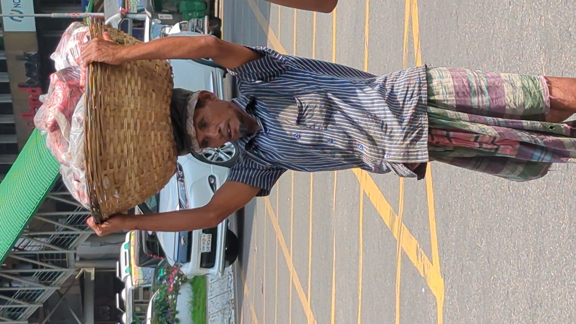 An elderly man carries a large, woven basket filled with goods on his head, standing on a street in Dhaka, Bangladesh. He is wearing a striped shirt, a colorful patterned lungi, and a cap. The street has yellow painted lane markings and vehicles parked on the side. A building with a green awning is visible in the background.