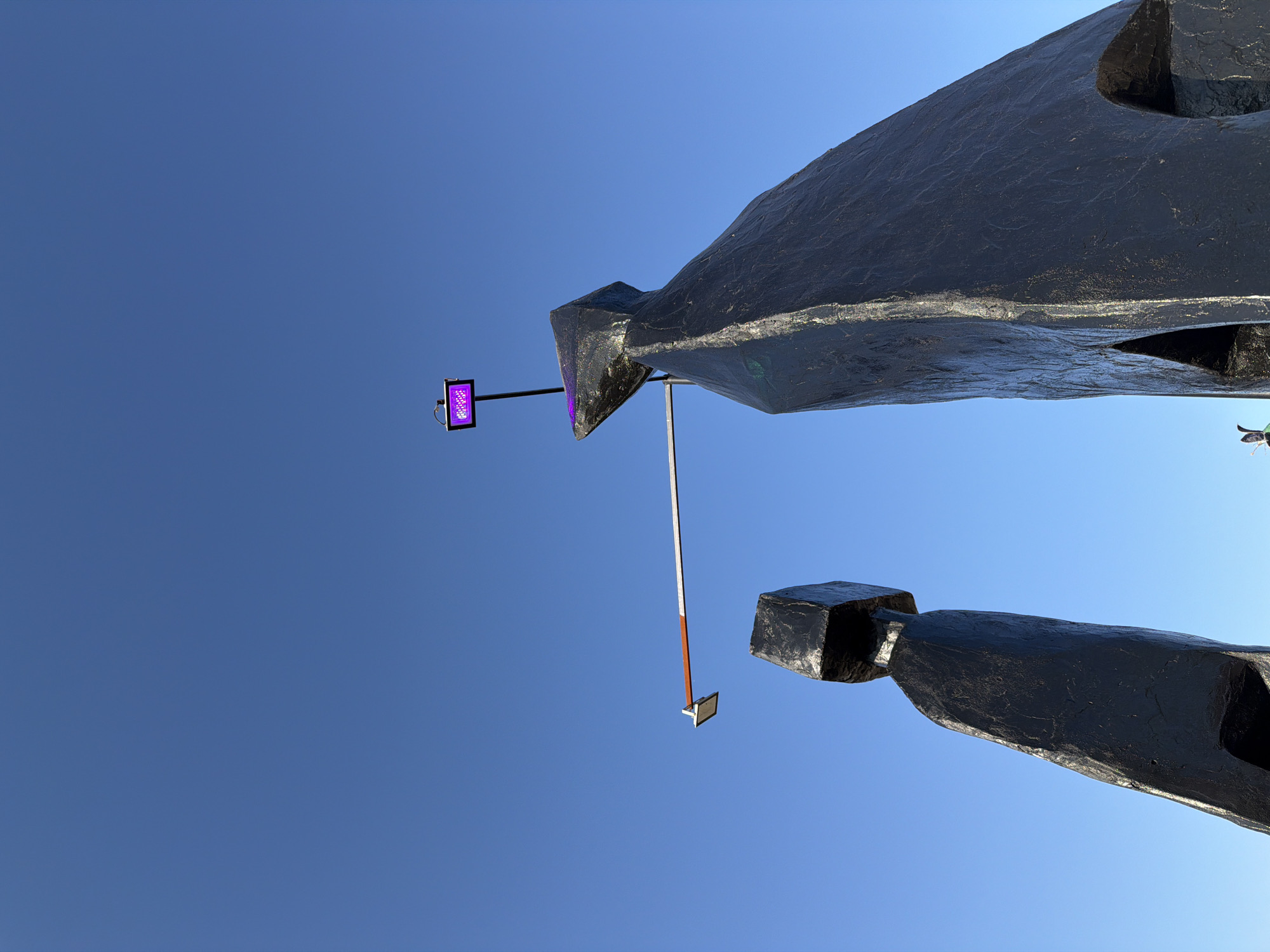 This is a low-angle, exterior, daylight shot of two abstract, dark-colored metal sculptures positioned against a clear blue sky. The sculpture on the left is shorter and has a block-like head. The sculpture on the right is taller and appears to have a more pointed head, with a section of purple light reflecting on its surface. Extending horizontally between the two sculptures is a slender metal rod with an orange band and a small rectangular metallic object at its end. Above and to the right of the taller sculpture, a small rectangular purple light fixture is attached to a thin black pole. The scene is located in Volzhsky, Russia, likely an outdoor public space.