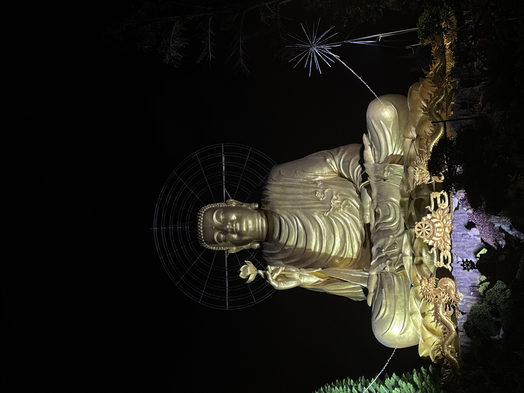 A golden statue of the Buddha stands illuminated against a dark night sky in Lâm Viên, Đà Lạt, Vietnam. The Buddha is seated in a meditative pose, holding a lotus flower. A decorative halo-like structure encircles the head. An ornate column adorned with dragons and other symbolic elements stands beside the statue. Nearby, foliage and other lighting elements are visible.
