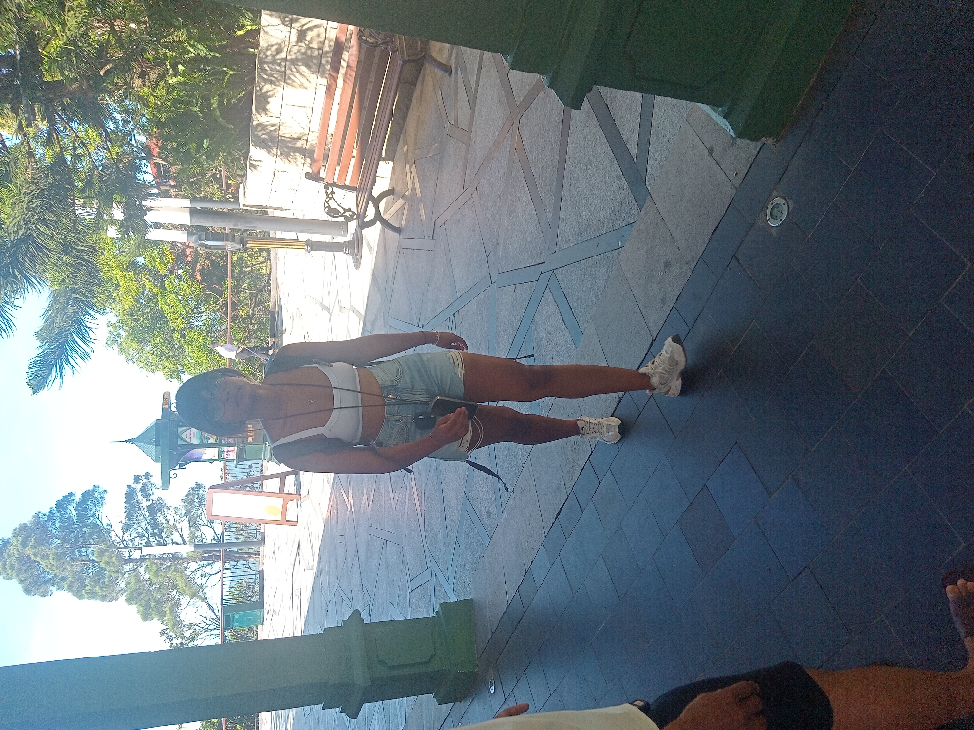 A woman walks towards the camera on a paved outdoor walkway. She is wearing a white halter top, denim shorts with ripped hems, and white sneakers. She has dark hair, glasses, and is carrying a dark backpack and a phone in her right hand. The walkway is paved with large gray tiles arranged in a geometric pattern. To the left, a large green pillar stands. In the background, there is a wooden bench, trees, and a clear sky. The scene appears to be in an outdoor public area, possibly a park or a tourist attraction. The lighting suggests it is daytime. This image does not contain any recognizable landmarks or features that would specifically place it in Đà Nẵng, Vietnam.