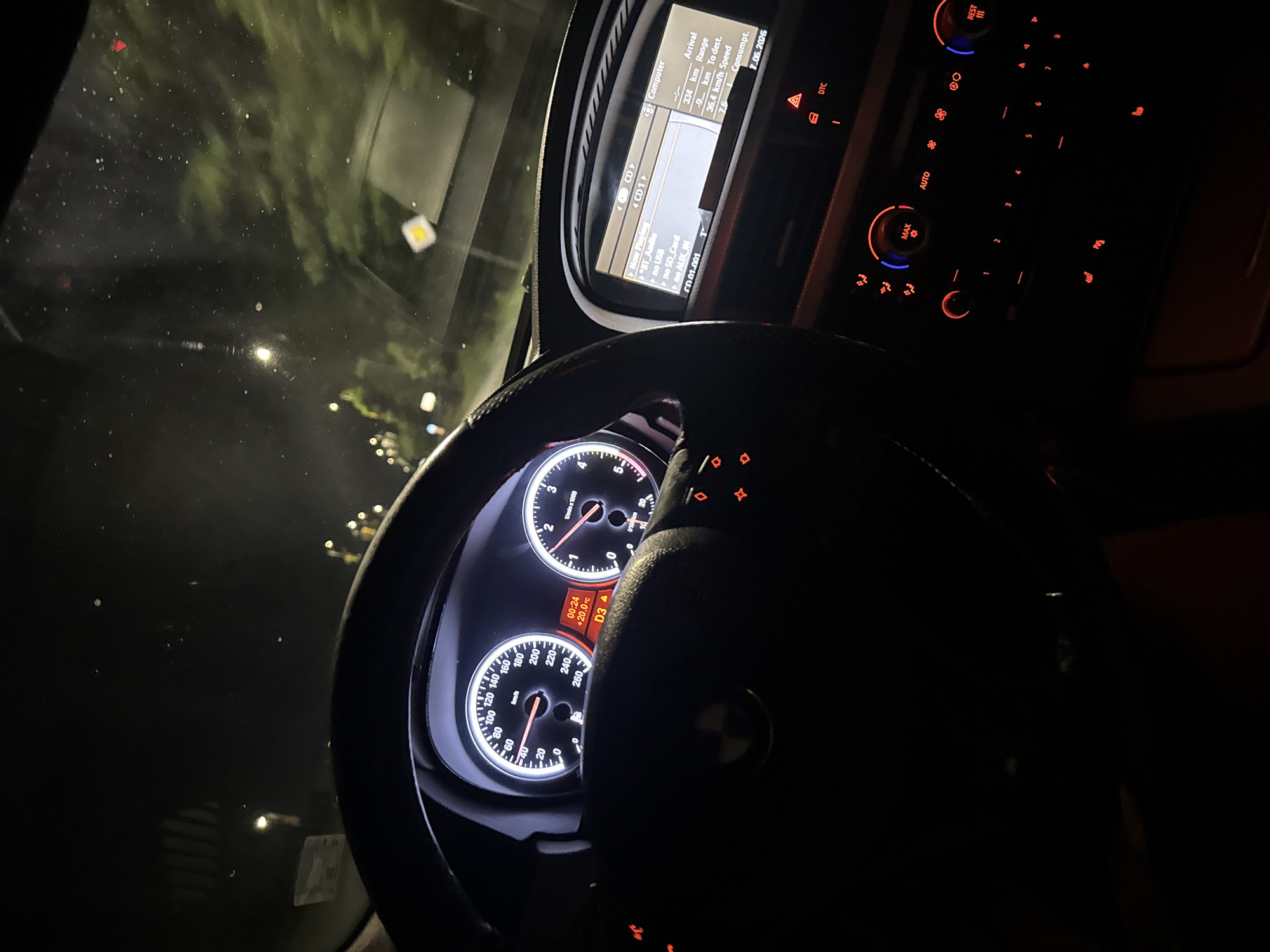 The image shows the interior of a car at night. The perspective is from the driver's seat, looking forward. The dashboard displays a speedometer, tachometer, and an infotainment screen. The speedometer indicates a speed of approximately 60 km/h. The infotainment screen shows "Now Playing CD" and a "Computer" display with information about arrival, range, speed, and fuel consumption. The center console features climate control buttons and knobs, illuminated in red and blue. Outside the car, trees and streetlights are visible through the windshield, suggesting a road in a city environment. The location is identified as Dimitrovgrad, Bulgaria.