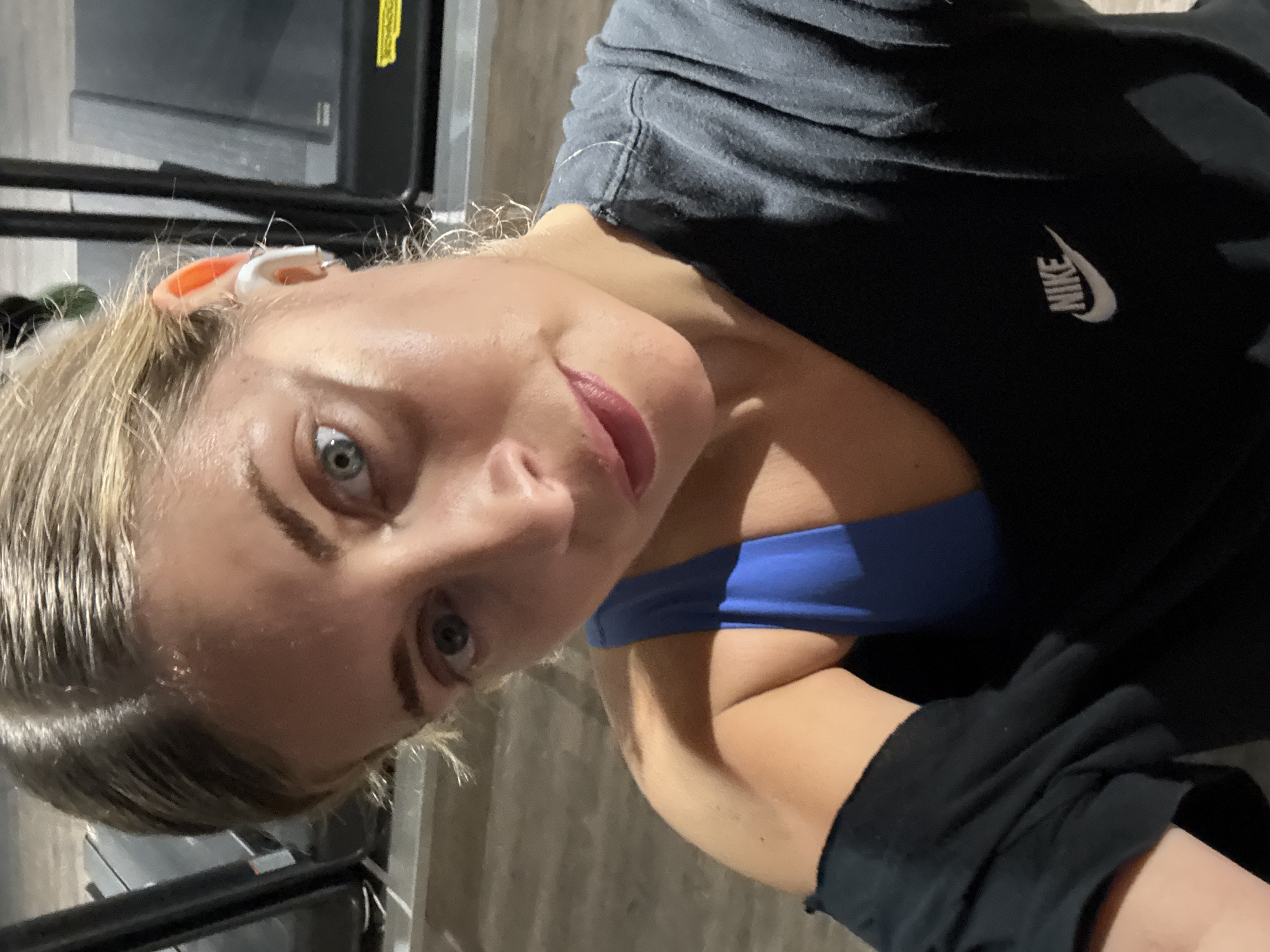 A close-up selfie, captured in an indoor setting in Paris, France, features a young woman looking directly into the camera. She has blonde hair and striking blue-grey eyes, with a neutral expression and lips tinted with a muted pink color. She is wearing a dark grey or black short-sleeved t-shirt that prominently displays a white Nike logo and swoosh on the chest. A bright blue sports bra or top is visible beneath her t-shirt. An orange and white earbud or hearing aid is noticeable in her right ear. The background is dimly lit, revealing dark, metallic structures that resemble gym equipment, along with a wood-paneled floor. The image suggests the woman is either during or after a workout, given her attire and the setting.