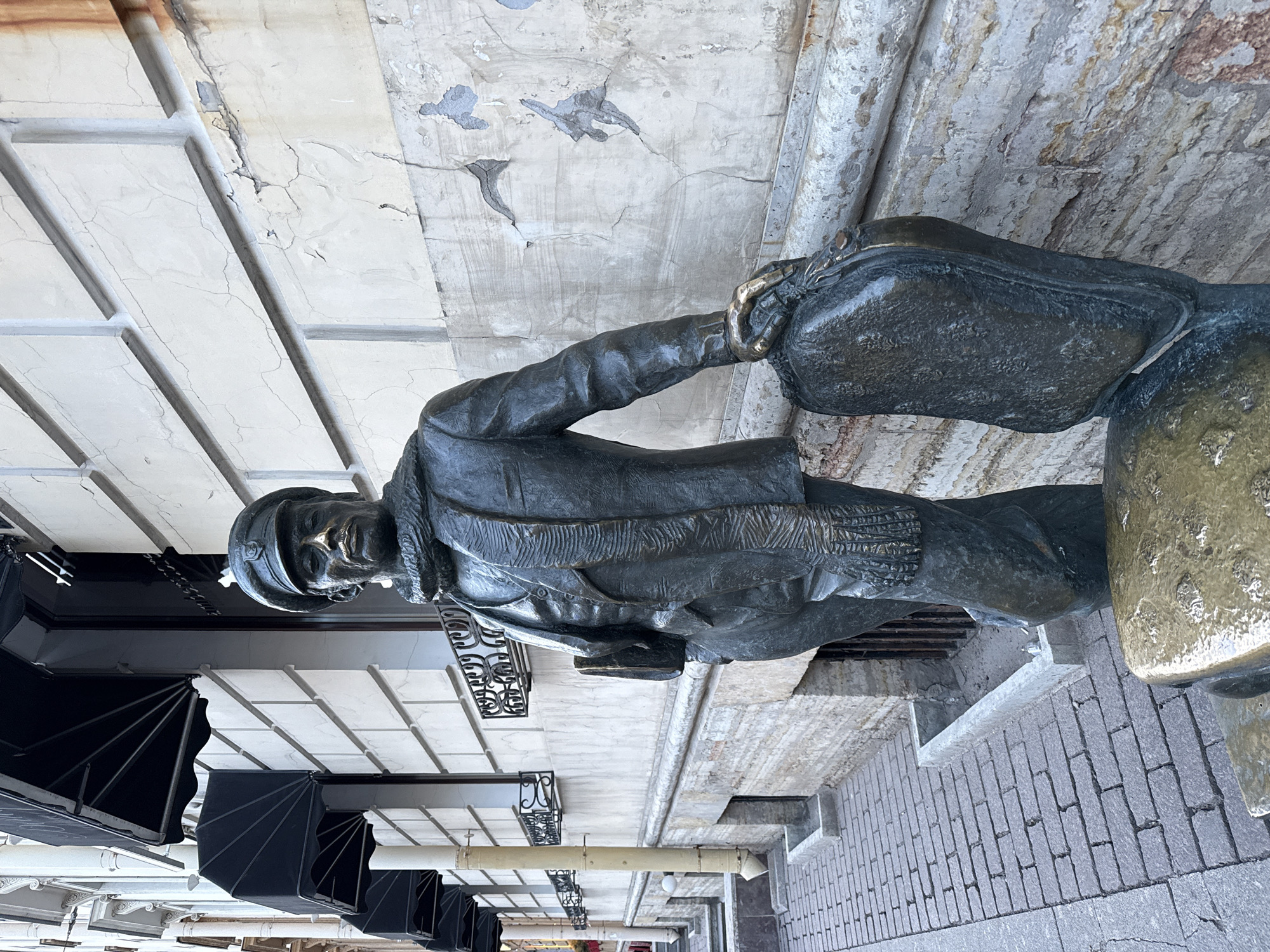 The image depicts a bronze sculpture of a man in period clothing, likely early 20th century, carrying a suitcase. He is wearing a cap, a jacket, and possibly a scarf. The sculpture is positioned on a brick walkway in what appears to be an urban environment. Above the sculpture, there is a building facade with a white and grey patterned surface, possibly indicating age and some disrepair, and black decorative awnings hanging from the building. There are other architectural elements in the background, including a railing and possible light fixtures. The environment suggests a historical area within the city of Saint Petersburg, Russia.
