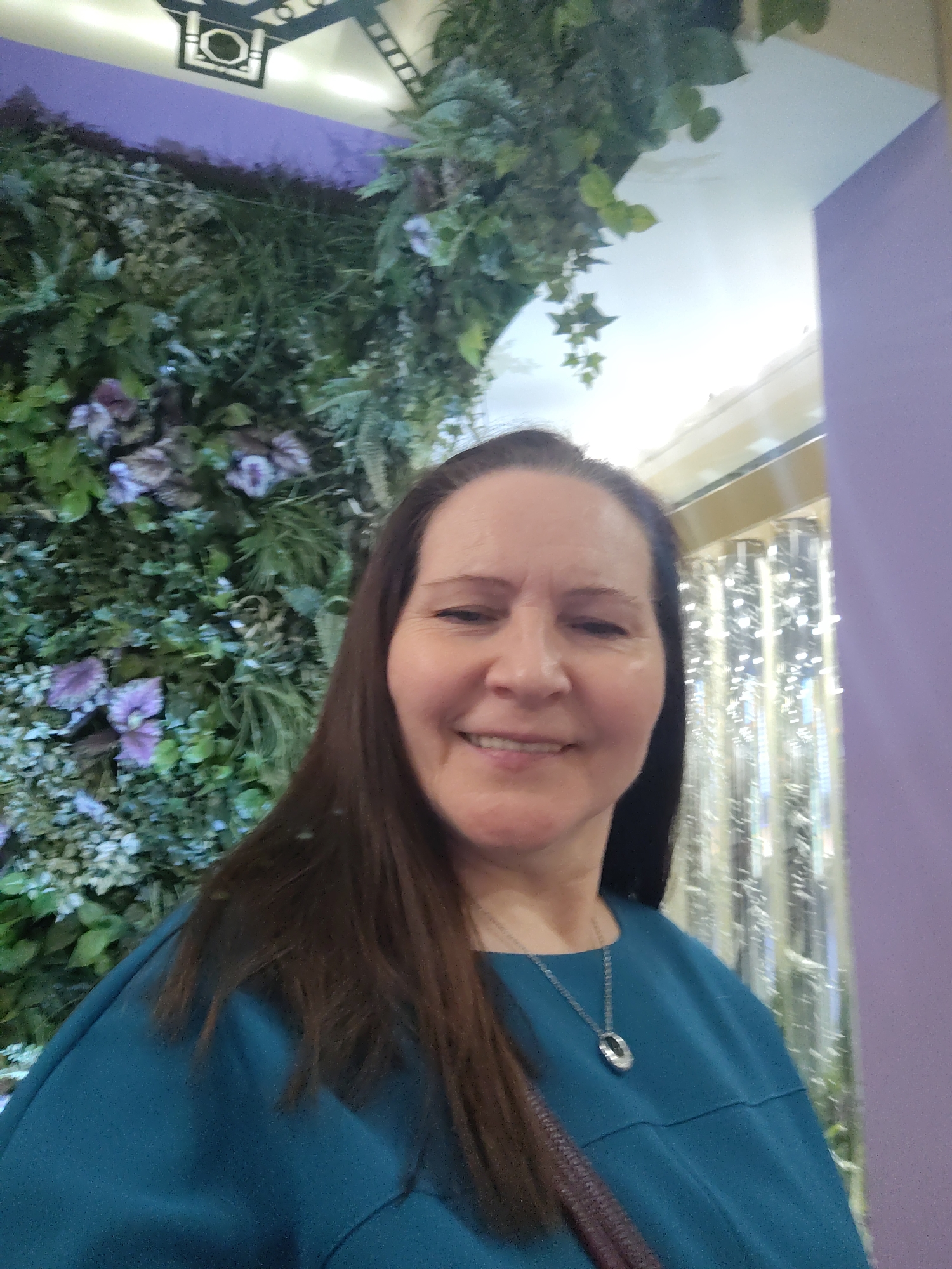 A smiling woman with long, dark brown hair is captured in a close-up, indoor, portrait-style photograph. She is looking directly at the camera, wearing a teal-colored top and a silver necklace with an oval-shaped pendant. The setting is an indoor decorative space, possibly in Moscow, Russia.
Behind the woman, to her left, is a prominent wall covered in lush artificial green foliage, interspersed with some purple or pink decorative leaves and flowers, resembling a vertical garden. Above this, a segment of a decorative ceiling feature with a dark geometric design is visible against a lighter ceiling. To her right, a smooth purple wall or partition is seen, behind which are shiny, reflective vertical strands, possibly part of a light installation or a decorative curtain, illuminated with bright spots. The woman's expression is joyful and relaxed, suggesting she is posing for the photograph. The lighting is soft and consistent with indoor artificial light. No text is visible in the image.