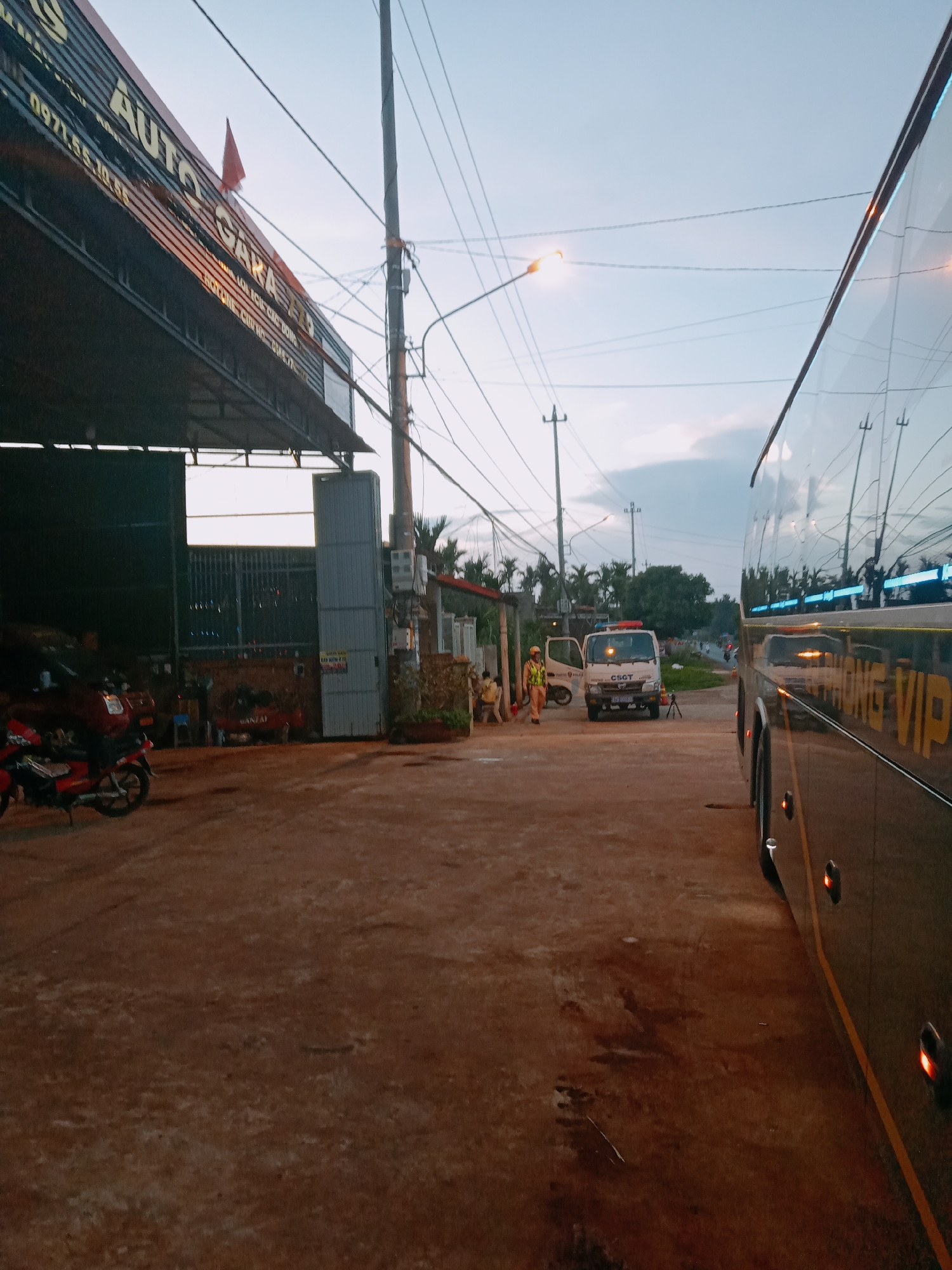 The image shows a street scene in Xuân Long, Vietnam. On the left is a building with a sign reading "AUTO GARA," under a metal awning. A red motorcycle is parked outside. Across the street, a white truck with a light bar on top and "CSCT" is parked. Two individuals wearing yellow reflective vests are standing near the truck. A large bus occupies the right side of the frame, with "VIP" visible on its side. Utility poles and power lines are strung overhead. The street appears unpaved and wet. The sky is overcast.