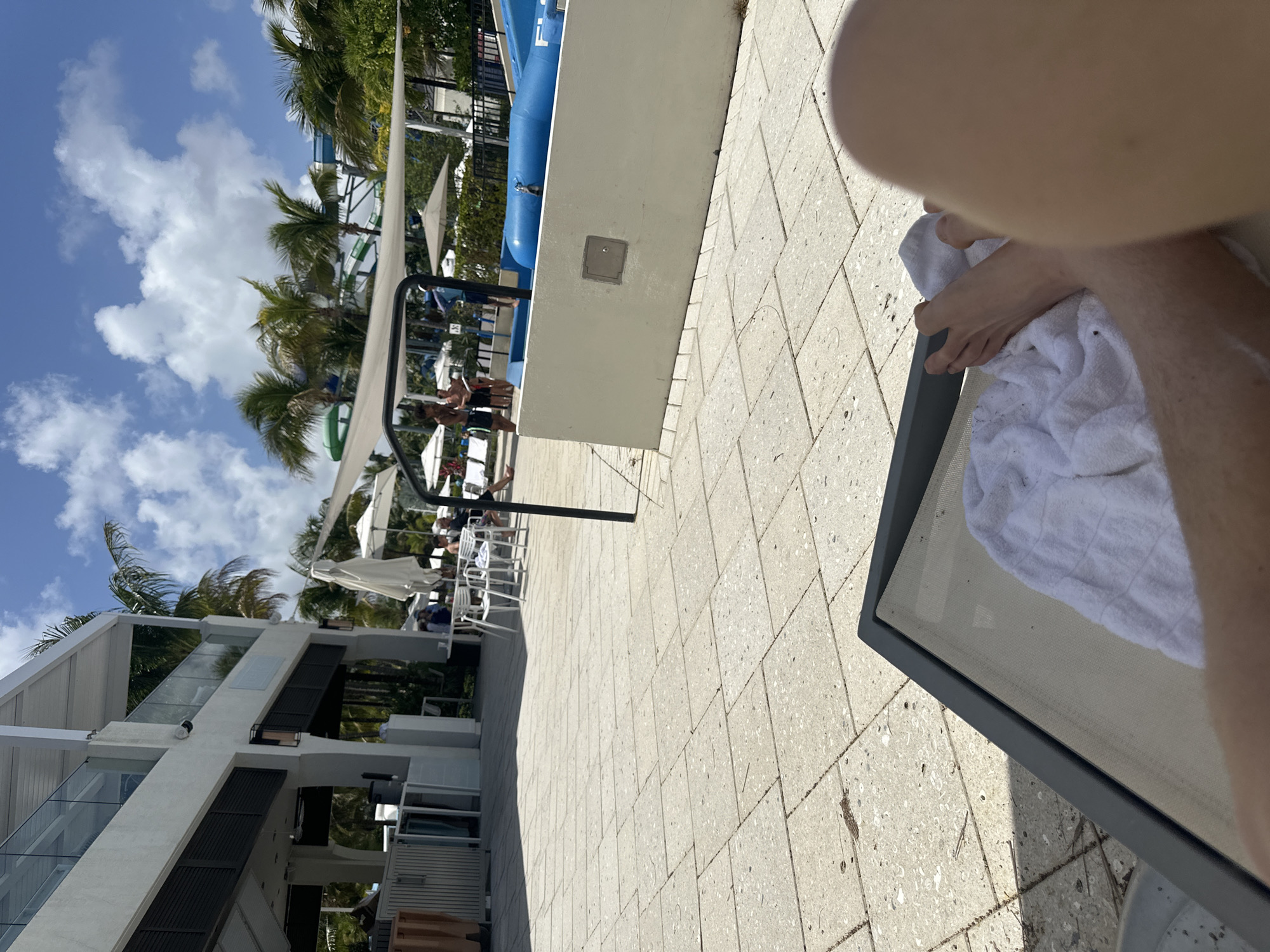 The image appears to be taken at a resort or pool area in Aventura, Florida, United States of America. The foreground features a lounge chair with a person relaxing, partially visible. The mid-ground shows other lounge chairs and umbrellas near a pool, with people sunbathing and enjoying the water activities. In the background, there are palm trees and buildings. The weather seems sunny, with blue skies and some scattered clouds. A structure that appears to be a water slide is also visible. The ground is tiled.