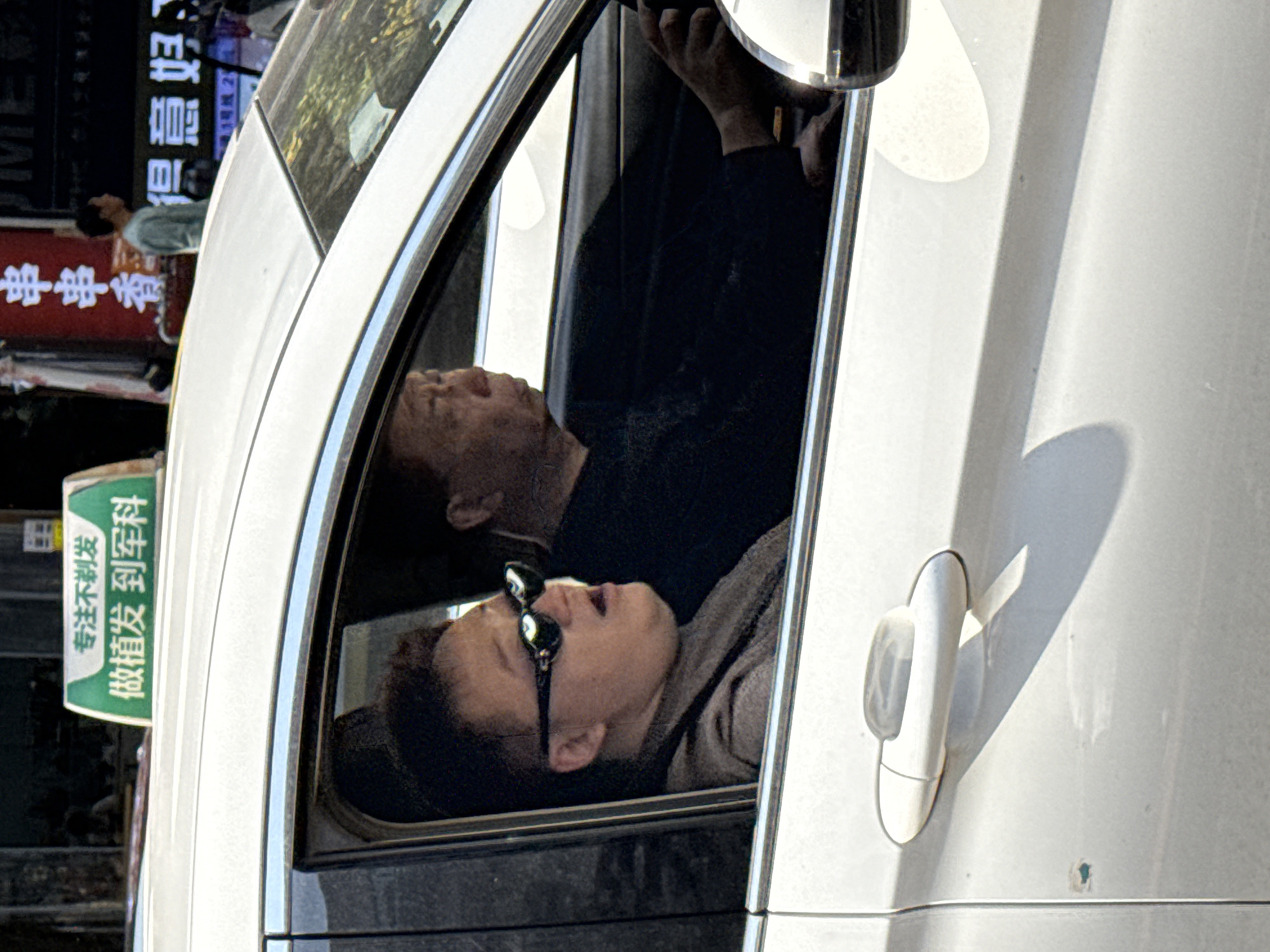 A high-angle outdoor shot captures the side of a white car on a sunny day in Yuzhong District, China. Inside the car, seen through an open window, are two individuals. A passenger in the foreground, wearing sunglasses and a brown top, has their head tilted back and mouth slightly open, appearing to look upwards or resting. Behind them, the driver, an older person with short hair, is also visible, with their head similarly tilted back, suggesting they are looking upwards or ahead.
Bright sunlight creates strong shadows on the car's white body and interior, highlighting reflections on the window and rearview mirror. The overall atmosphere suggests a casual moment, perhaps while waiting in traffic or parked.
In the blurred background, several Chinese street signs are visible. A green sign advertises a hair transplant service, reading "专注不剃发 做植发到军科" (Specializing in no-shave hair transplants at Junke). Further back, a red sign shows "串串香" (Chuan Chuan Xiang), indicating a food establishment. Another sign includes "MED," "得意好" (Deyih-Hao), and "通1号线 24" (Pass Line 1, 24), likely pointing to a subway line and exit. The urban setting is active, with hints of buildings and other elements in the distant background.
