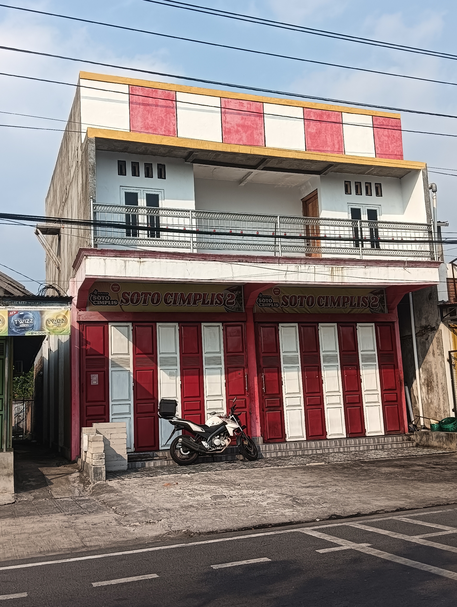 The image shows a two-story building with a red and white checkered pattern on the upper facade and a yellow trim. The lower facade is painted red and white, with the text "SOTO CIMPLIS 2" prominently displayed. There are multiple red and white shuttered doors on the ground floor. A white motorcycle with a black storage box is parked in front of the building. The scene appears to be on a street in Sragen, Indonesia, with power lines visible against a blue sky.