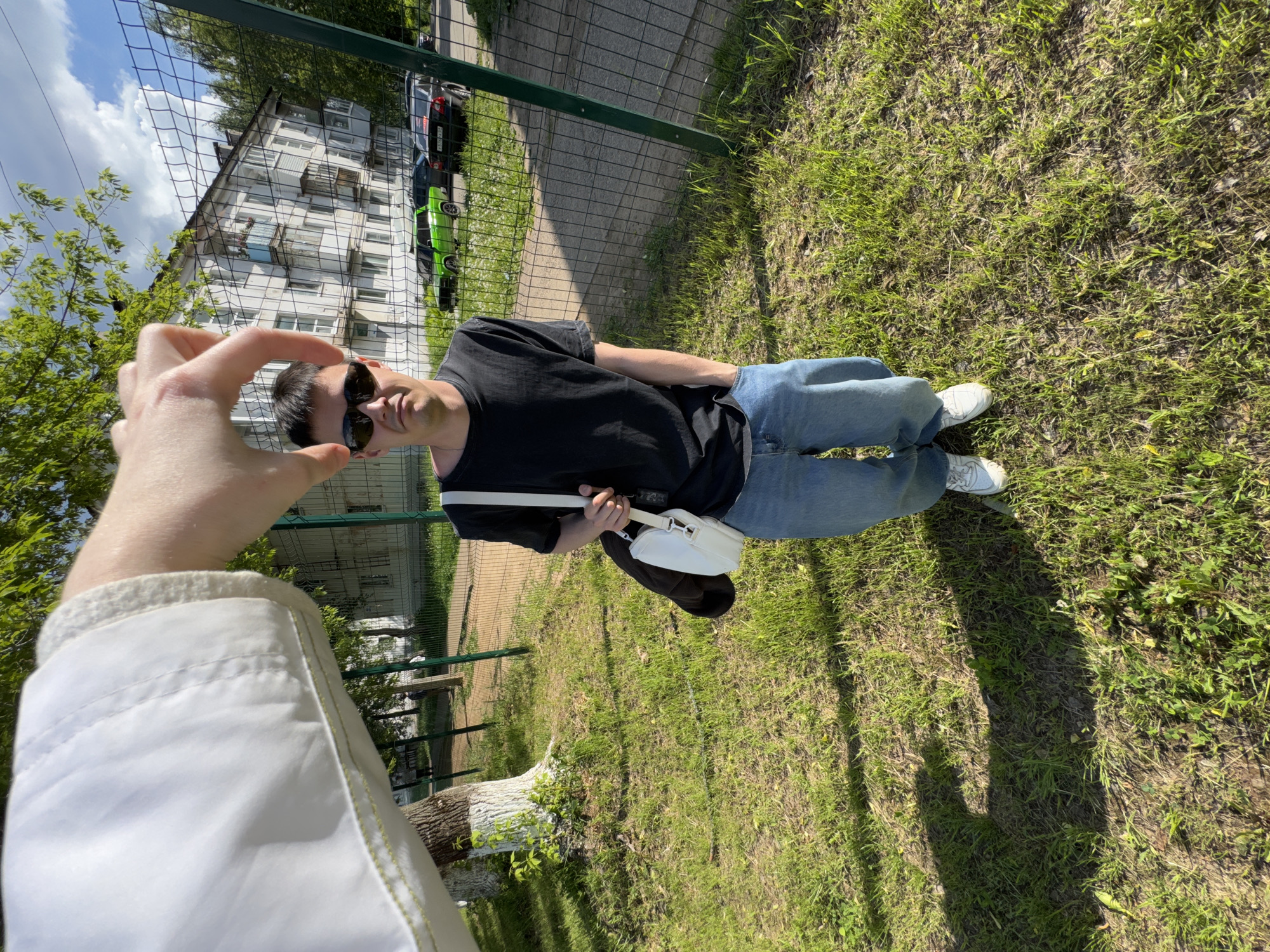 The image captures a sunny outdoor scene, likely in Chaikovsky, Russia, given the Cyrillic lettering on vehicles visible behind a chain-link fence and residential buildings in the background. A person is being photographed. The photograph is oriented vertically.
The focal point of the image is a man standing on a grassy area. He is wearing a black t-shirt, blue jeans, white sneakers, and sunglasses. A white shoulder bag hangs across his body.
In the foreground, a person's arm and hand are visible, with the fingers forming a circle around the man's head, suggesting they are part of taking the photograph. The photographer wears a white jacket.
The background includes a building with several stories and windows, a chain-link fence, parked cars, and green trees. The setting appears to be a park or a green space adjacent to a residential area.