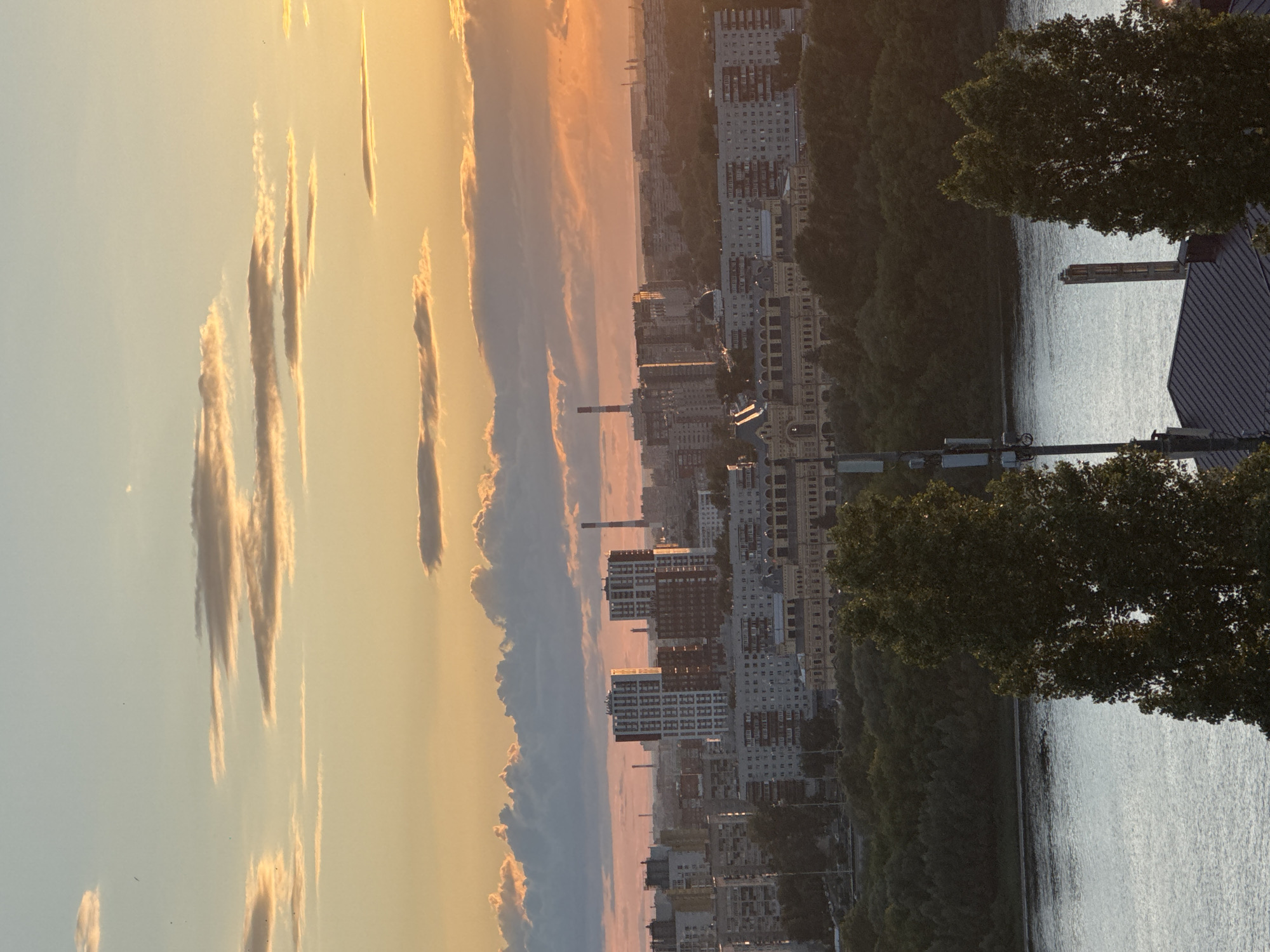 This is a wide, eye-level shot of the Nizhny Novgorod cityscape at sunset, with a river in the foreground and a warm, orange and pink sky filled with scattered clouds. The city is densely packed with apartment buildings of varying heights and architectural styles, ranging from older, grey structures to more modern, taller buildings with patterned facades. Some prominent landmarks are visible, including two tall, rectangular buildings with dark and white grid patterns, and a tall, red and white-striped industrial chimney on the left side of the image. A large, yellow building with arched windows and a dome structure is visible in the middle ground. A wide, calm river flows across the bottom of the frame, reflecting the sky's colors. Lush green trees line the riverbank, creating a natural buffer between the water and the urban sprawl. In the foreground, partially obscuring the view of the river, are the tops of two large, dark green trees, with a tall cell tower with multiple antennas to the left of center. A dark, sloped roof of a building is also visible in the lower right corner, and a section of a grey, brick structure with a narrow, tall window opening is visible to the right of the cell tower. There are no people visible in the image. The lighting suggests it is either early morning or late evening, with the sun low on the horizon casting a warm glow.