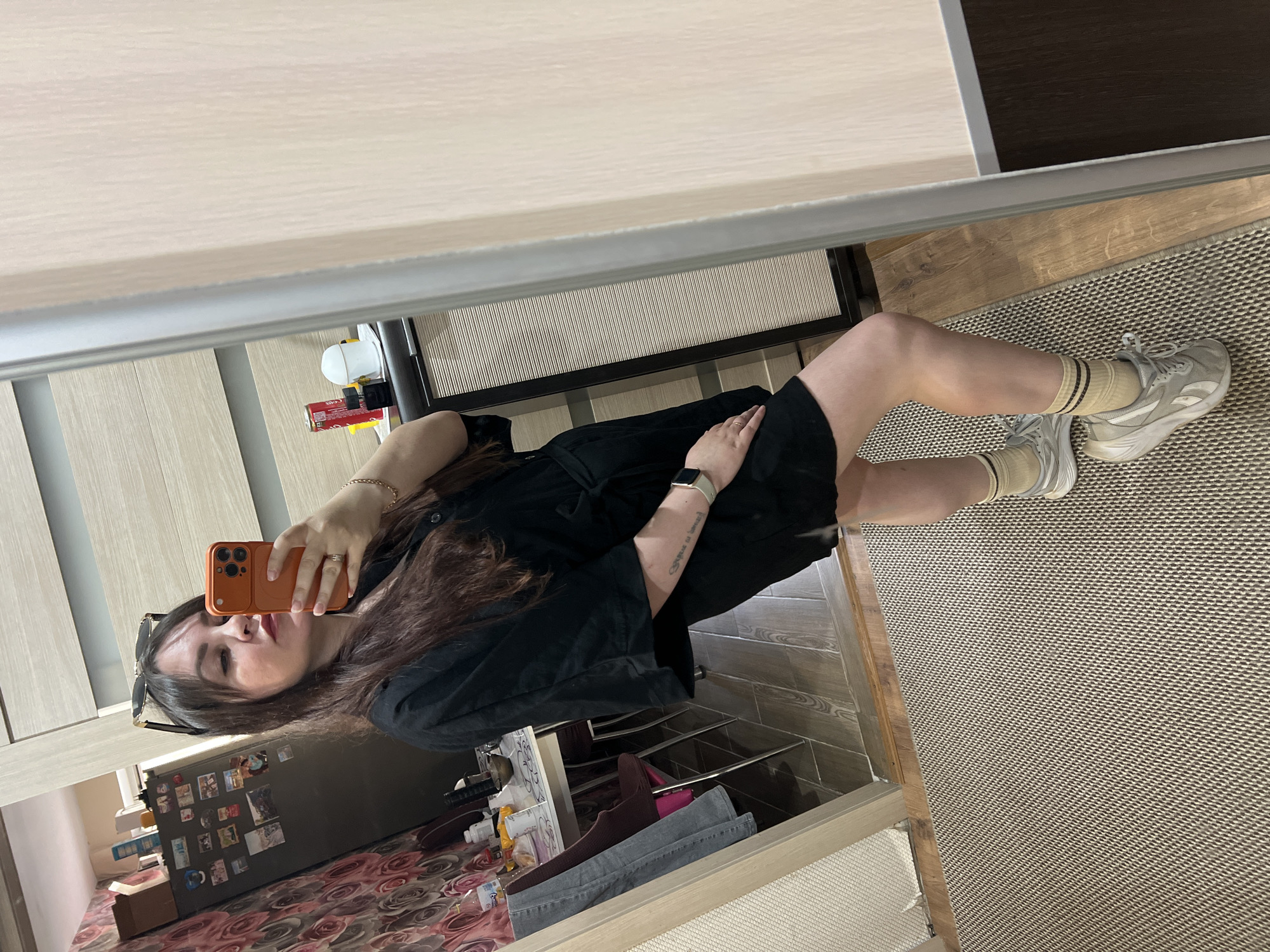 A person takes a selfie in front of a mirror. The person is wearing a black dress, beige socks with stripes, and grey sneakers. The mirror reflects a room with a refrigerator covered in magnets and a wardrobe. A rug is on the floor. The location is within a building in Noviy, Russia.