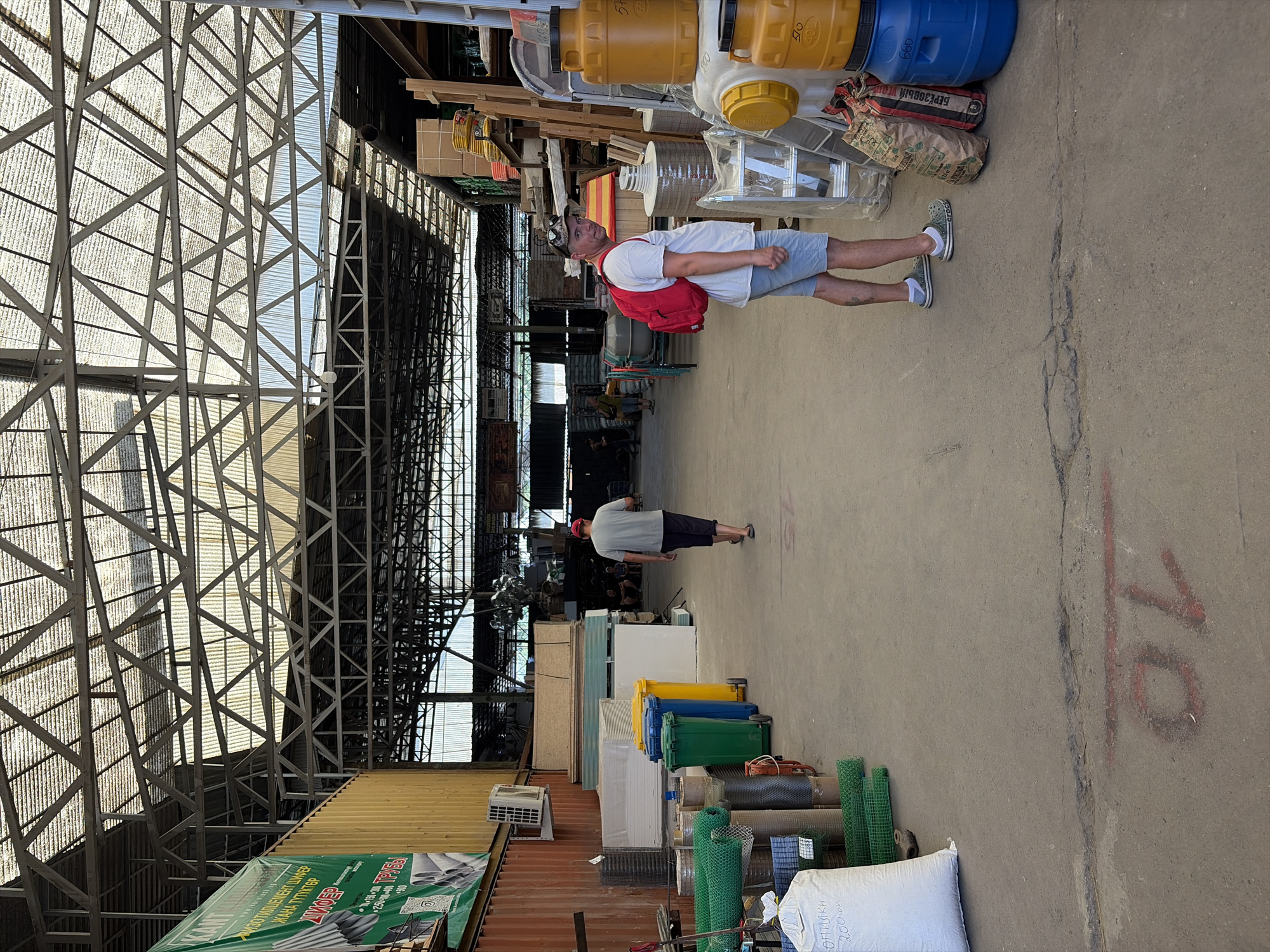 The image depicts an outdoor market setting in Kant, Kyrgyzstan, characterized by a large, open-air structure with a metal roof supported by a truss system. Sunlight streams through the corrugated roof panels, creating patterns of light and shadow on the concrete ground.
In the foreground, a man wearing a white t-shirt, blue shorts, and camouflage-patterned glasses on his head, with a red backpack slung over his left shoulder, is looking towards the camera. He is standing next to stacks of large, yellow and blue plastic barrels, as well as bags of materials.
Further into the scene, another man in a gray t-shirt and dark shorts is walking away from the camera, towards the background of the market. Several colorful plastic trash bins (green, blue, and yellow) are visible in the middle ground. A variety of goods, including rolls of mesh fencing, white sacks of material, and what appear to be construction boards, are displayed throughout the market. In the distance, other shoppers and market stalls can be observed. The overall atmosphere is that of a bustling, open-air marketplace with a wide array of goods.