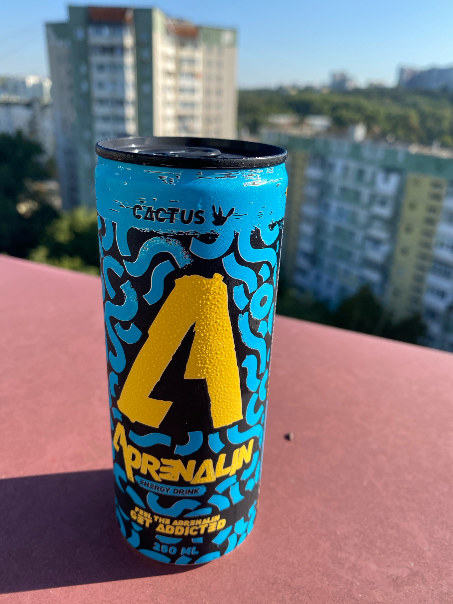 A close-up, eye-level, vertical shot captures an Adrenaline Cactus energy drink can against a blurred urban backdrop. The can, predominantly blue with black and yellow graphic elements, stands on a reddish-brown surface, likely a balcony railing. Condensation is visible on the can, suggesting it is cold. The background features out-of-focus apartment buildings, characteristic of Chișinău's residential areas, with greenery and a clear blue sky. The lighting suggests a sunny day. The text "ADRENALIN ENERGY DRINK" is clearly legible on the can, as well as "CACTUS" and "250 ML".