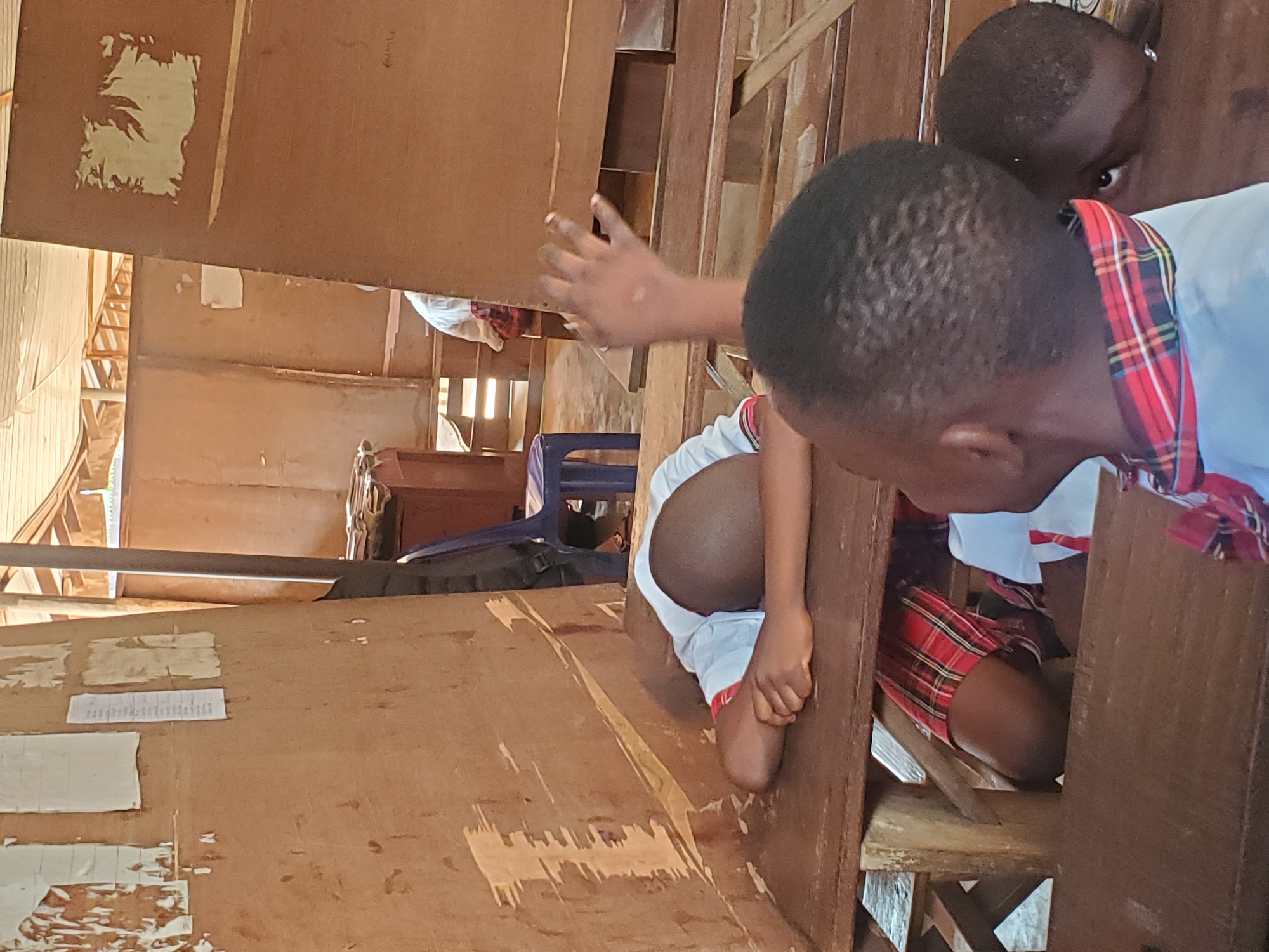 Two children are in what appears to be a wooden church or similar building. One child is kneeling in a pew, and another is either standing next to the pew. One child is wearing a white shirt and plaid skirt. The specific location is Ukwu-Abwa, Nigeria. The environment is a simple wooden structure.