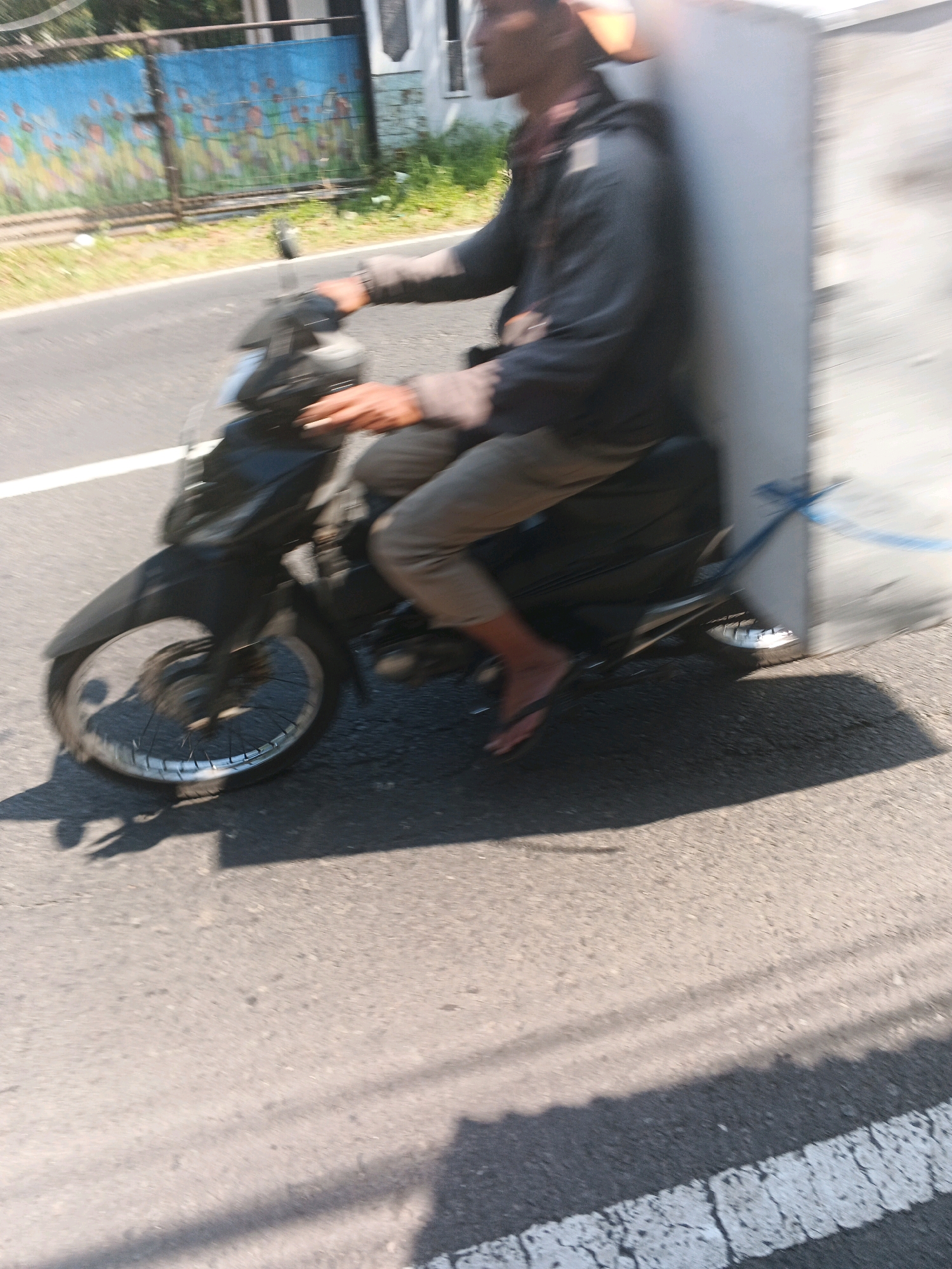In Bandungan, Indonesia, a person is riding a motorcycle on a street. The motorcycle is black and has a silver rim on the front wheel. The rider is wearing a dark jacket, tan pants, and flip-flops. They are carrying a large, light-colored rectangular object attached to the back of the motorcycle, secured with a blue rope. The background shows a blue fence with colorful artwork, green vegetation, and buildings. The scene is captured with motion blur, suggesting the motorcycle is in motion. The road is asphalt with a white line marking. Shadows of the motorcycle and rider are cast on the road.