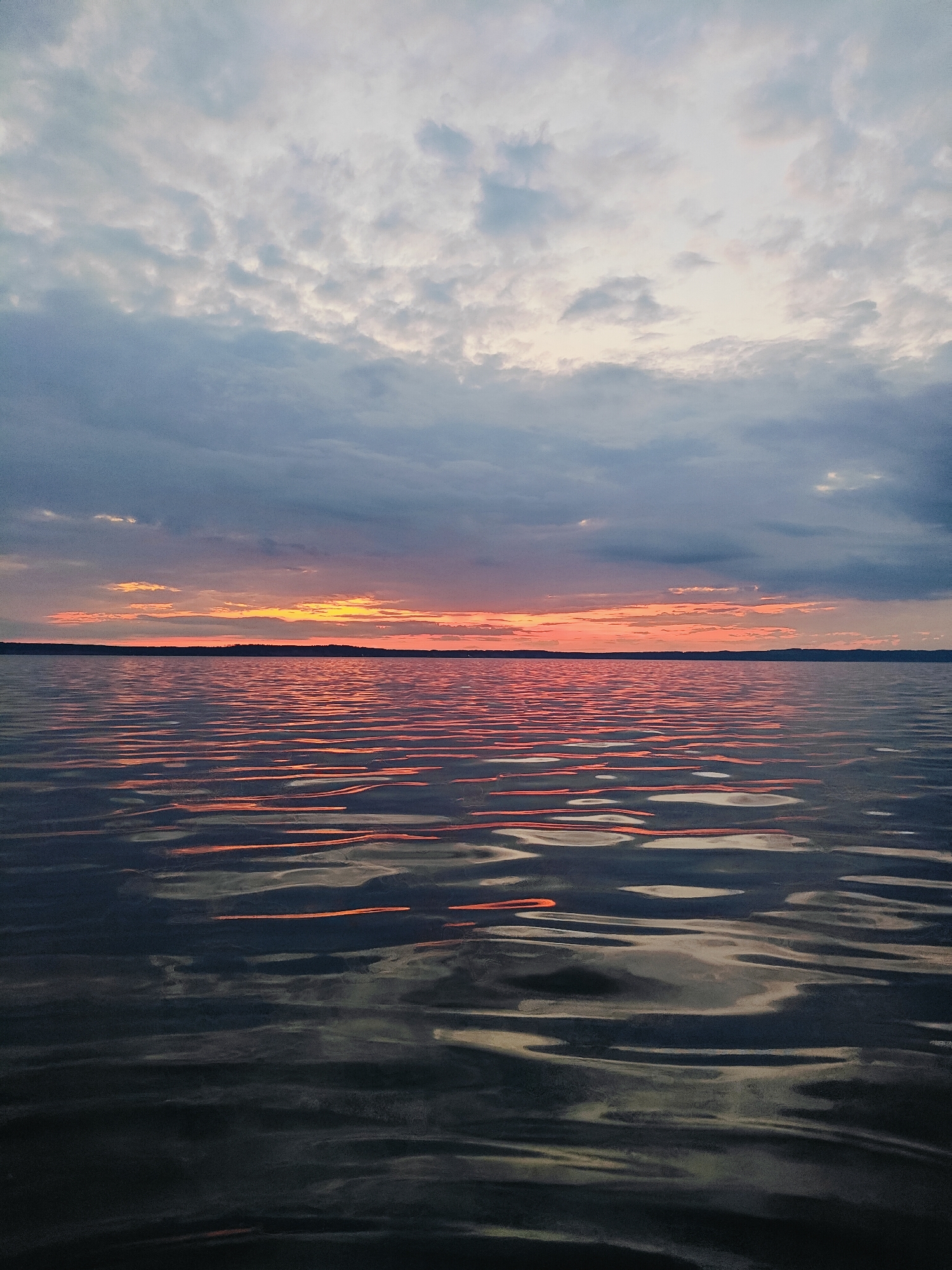 A sunset over a body of water is depicted in the image. The sky is filled with clouds, ranging in color from dark gray to light pink and orange near the horizon. The water is rippled, reflecting the colors of the sunset. A dark, distant shoreline is visible along the horizon. The scene is serene and peaceful. The image was taken in Chaykovsky, Russia.