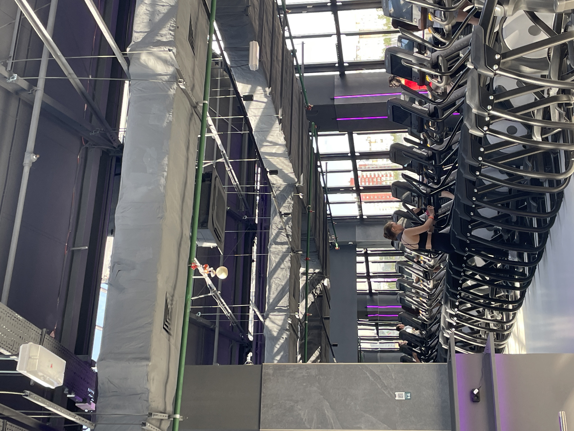 The image captures an indoor scene at a gym in Kazan, Russia. A row of stationary bikes dominates the right side of the frame, with individuals exercising on them. The building's architecture features exposed ductwork, scaffolding, and structural elements, giving an industrial aesthetic. A large duct, wrapped in a protective covering, runs vertically. The background reveals a grid-like window structure offering a view of the cityscape. The lighting is bright, likely due to natural light from the large windows.