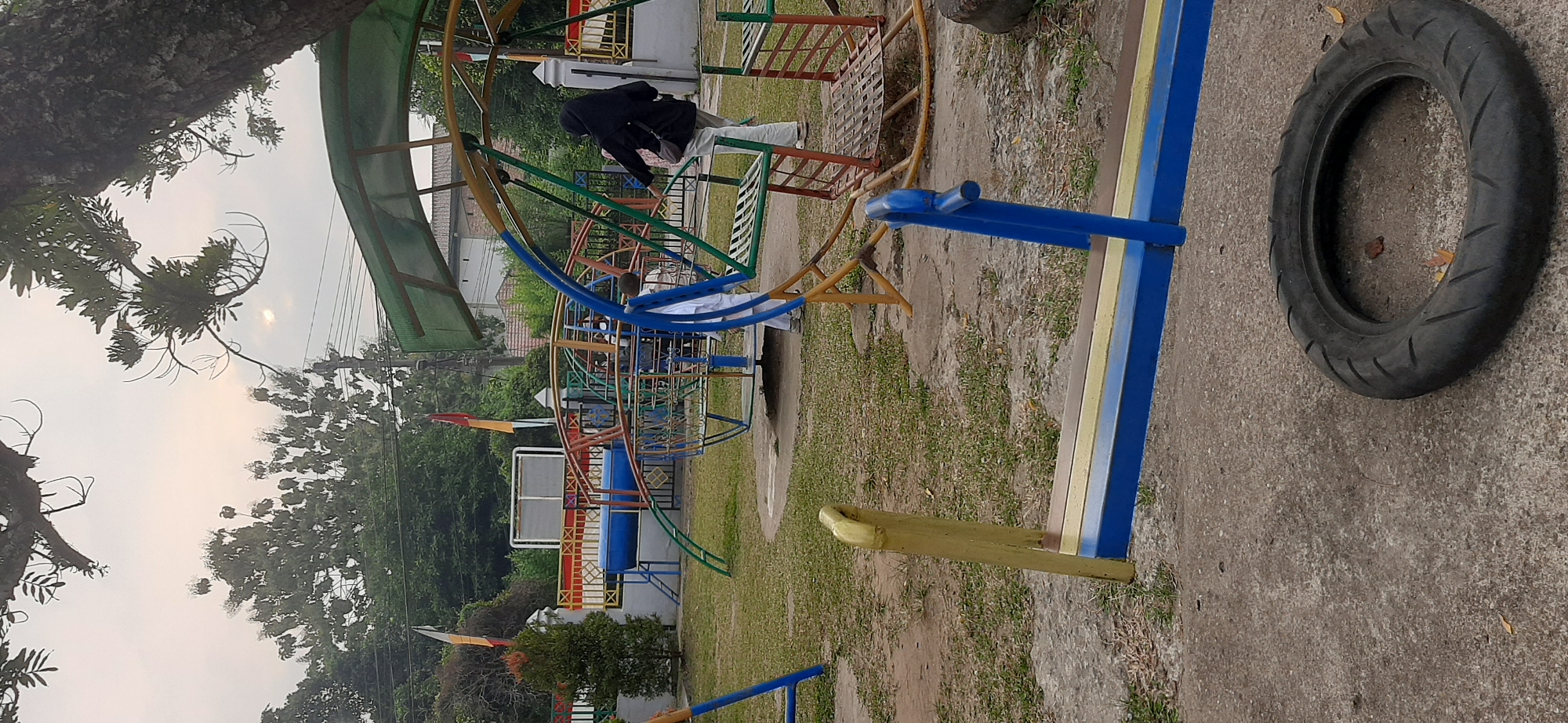 The image depicts an outdoor playground, likely in Jambi, Indonesia, as indicated by the presence of Indonesian flags and the general environment. The playground features a variety of colorful metal and plastic equipment, including a seesaw in the foreground, a swing set, and a larger, multi-component climbing structure in the middle ground. A person wearing a black hijab and white pants is visible interacting with the playground equipment, specifically near the swings. Another child in a white outfit is also present. The ground is a mixture of grass and concrete. The sky is overcast, suggesting it may be early morning or late afternoon. A large tree with prominent branches partially obscures the top right portion of the image, casting shadows over the scene. In the background, a low wall with decorative elements and some vegetation can be seen, along with buildings and utility poles. A discarded tire lies on the concrete surface in the lower right corner.