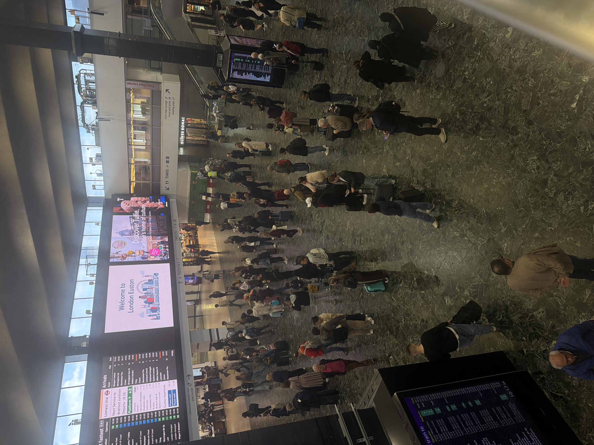 The image shows an overhead view of a crowded concourse inside London Euston railway station in the city of London, United Kingdom. Many travelers with luggage wait for trains. Digital display boards show arrival and departure information, including services to destinations like Watford Junction, Manchester Picc, and Wigan N Western. Advertisements indicate nearby attractions, such as the "Tullie, Carlisle's home of culture" and the "First Class Lounge," with signs for amenities like toilets, platforms, and a Caledonian Sleeper Club guest lounge. People are looking at the boards for train schedules.