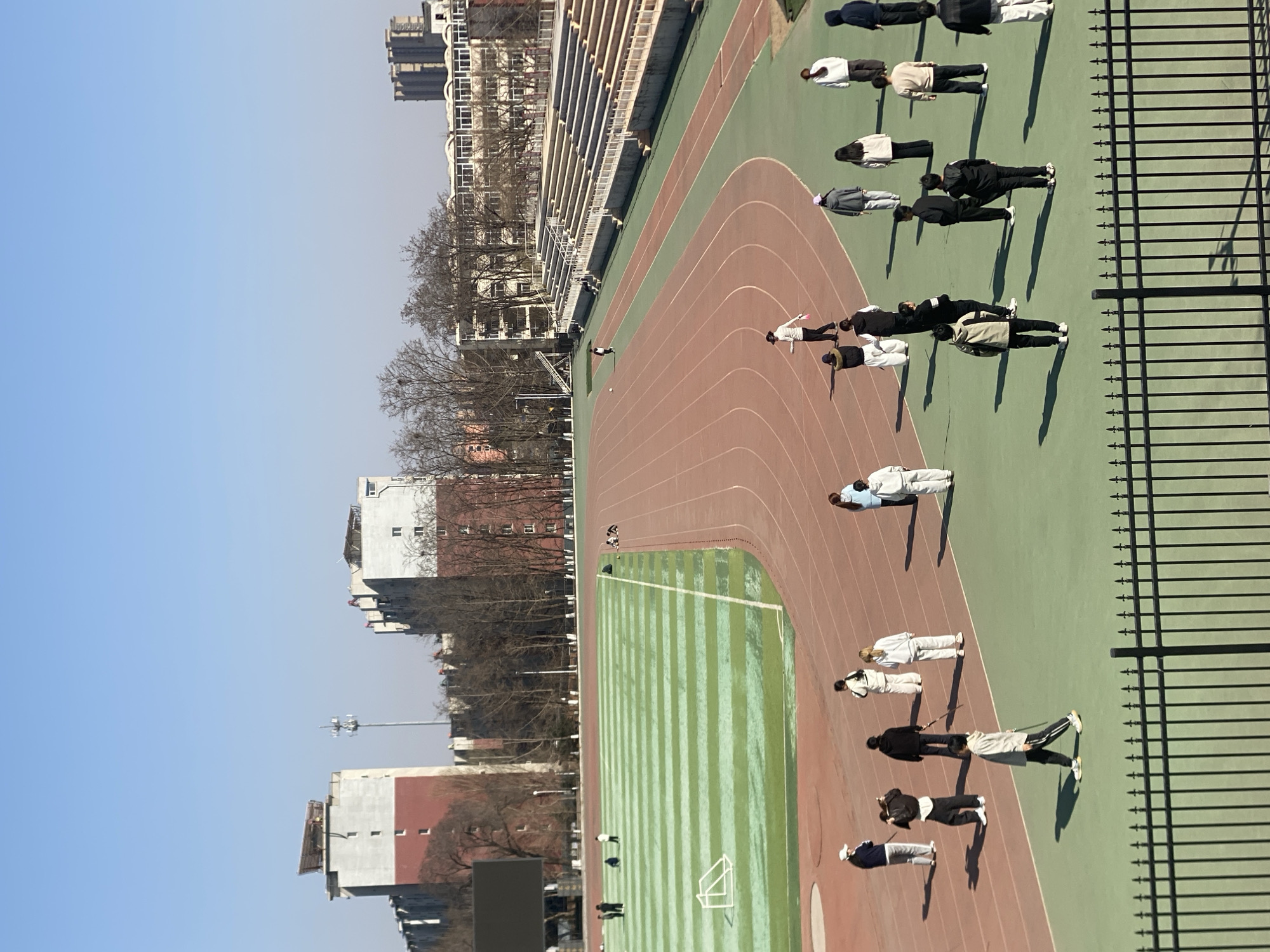 The image shows an elevated view of an athletic field and track in Zhongtun, China. A group of approximately a dozen people are spaced apart, lying on the green infield near the track lanes. The track itself is a reddish-brown color with painted lanes. A tall metal fence runs parallel to the track on the right side of the image. Behind the athletic field are rows of buildings, including low-rise apartments and a taller, possibly residential, building in the background. The sky is clear and blue. Shadows cast by the people suggest the sun is at an angle, indicating it is either morning or afternoon. The overall scene is one of an organized activity, likely some form of physical exercise or training, taking place on the athletic field.