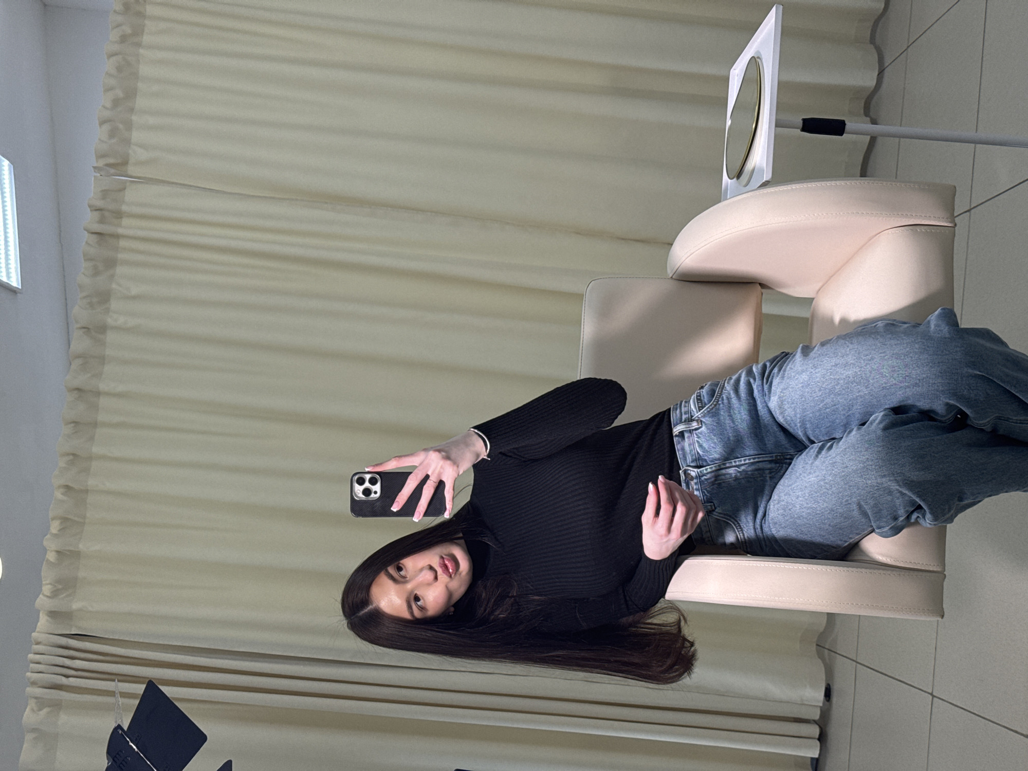 A woman is reclining in a salon chair, holding a black smartphone to take a selfie. She is wearing a black turtleneck sweater and blue jeans. Her long, dark hair is spread across the chair.
The chair is beige and appears to be in a salon setting. A large curtain is behind the chair and a makeup mirror is beside it. The image was taken indoors. The location is identified as the city of Chaikovsky, Russia.