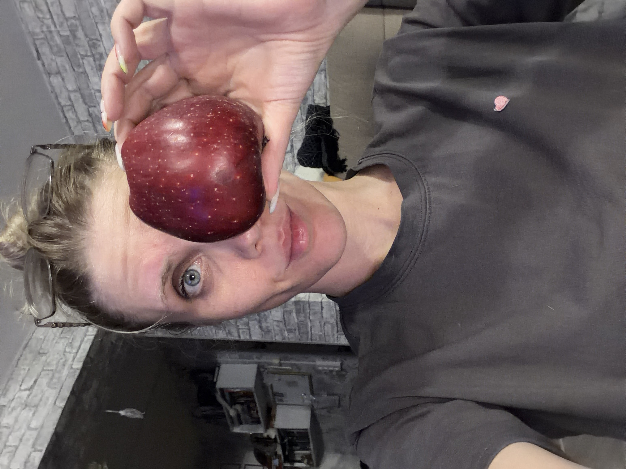 A woman with light skin and blue eyes is holding a red apple in front of her right eye. She has blonde hair pulled back in a bun and is wearing glasses perched on her head. She is wearing a dark gray t-shirt with a small red heart design on the upper right chest. She is holding the apple with her right hand, which has long, painted fingernails. The background includes a gray brick wall and a storage unit. The location is not specified to be in Chaikovsky, Russia.