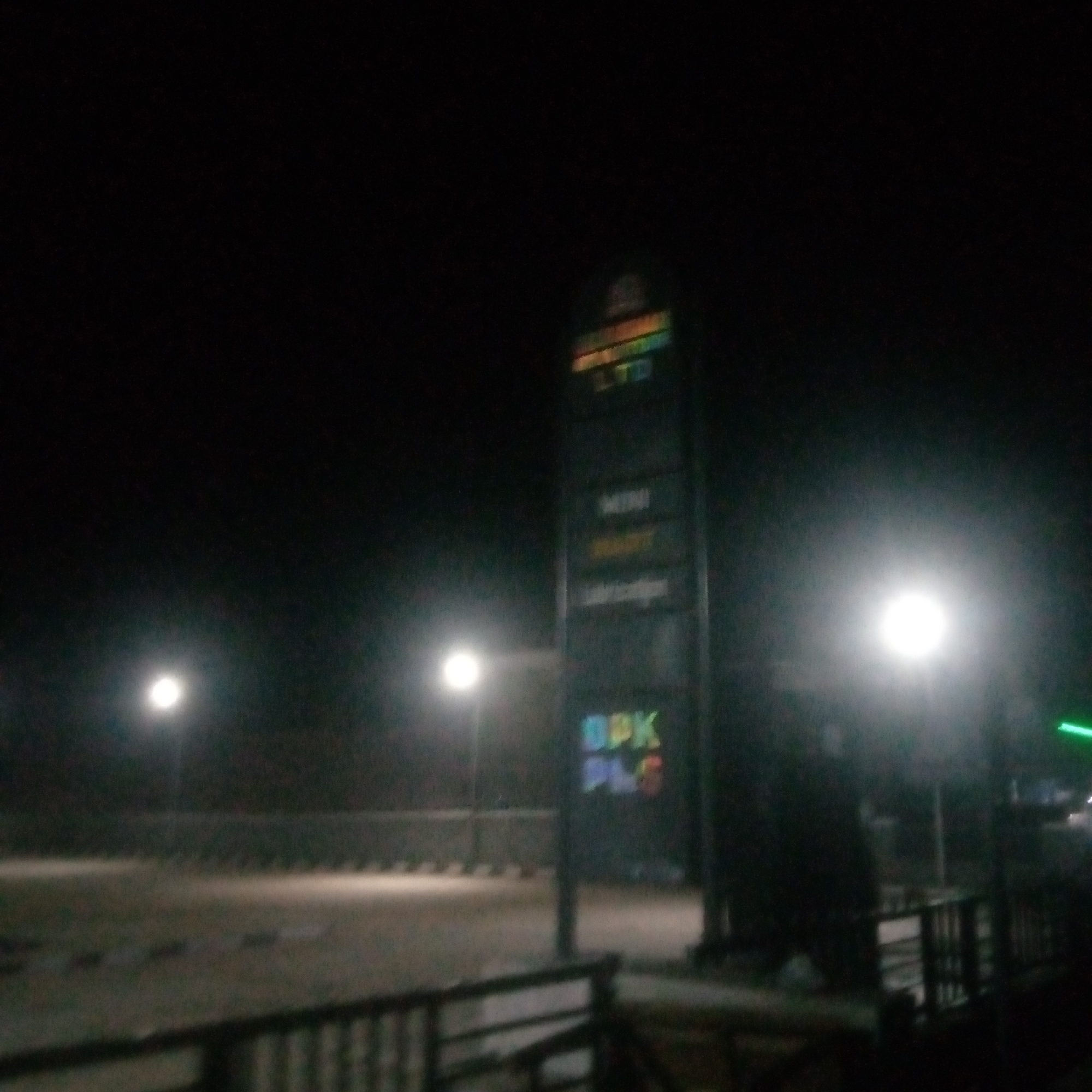 A tall, illuminated signpost stands in the foreground, displaying various words in different colors, possibly indicating businesses or services. The sign is lit, making the text visible against the dark night sky. To the left of the sign, a street light illuminates a paved area and a low barrier, suggesting a road or walkway. The background is dark and indistinct, with faint lights visible in the distance. The overall scene appears to be an outdoor urban environment at night.