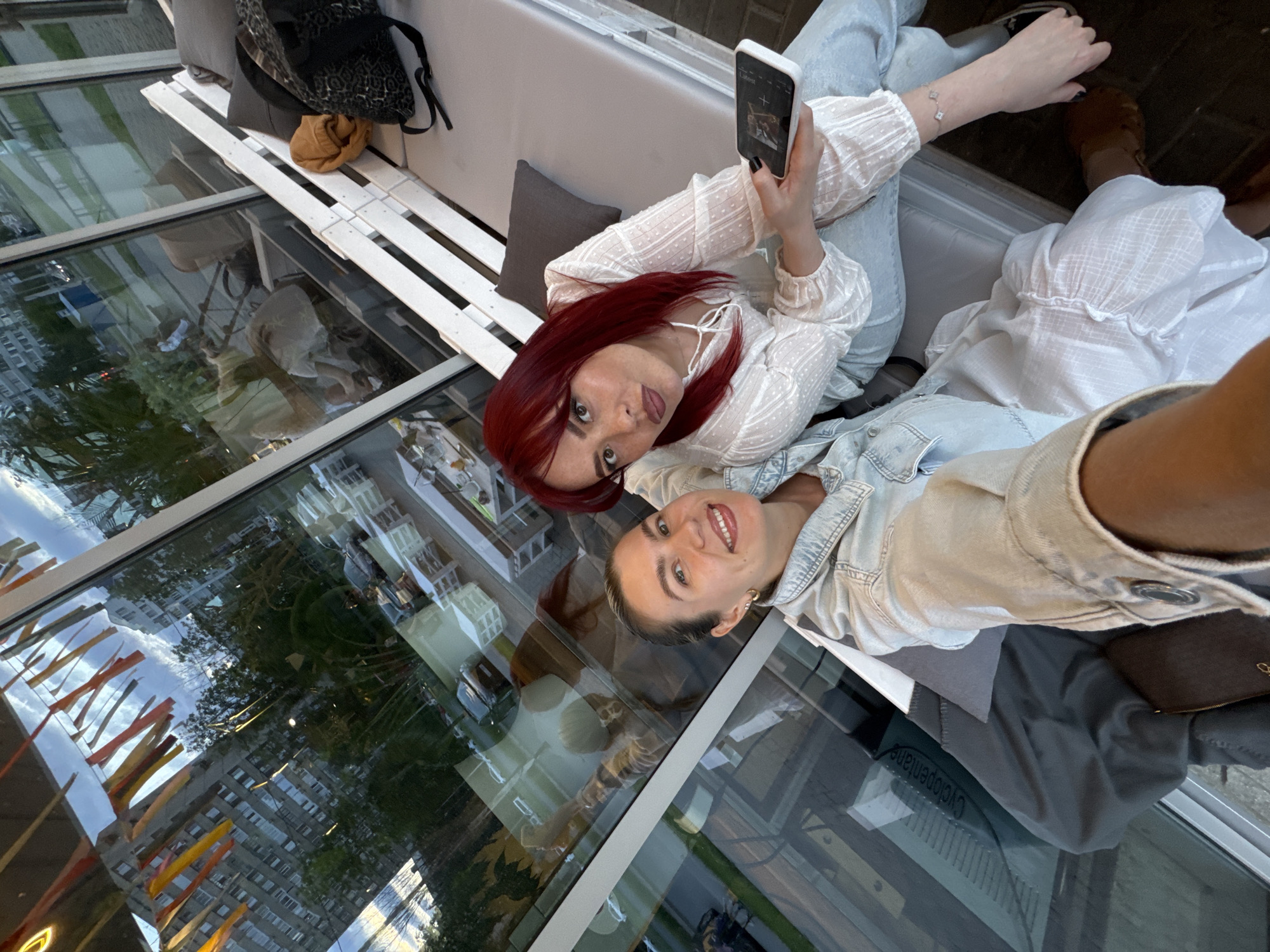 Two women are seated on a cushioned bench inside a cafe with large windows. The woman on the left is wearing a denim jacket and a white dress. She has her hair pulled back in a ponytail and is smiling at the camera. The woman on the right has vibrant red hair and is wearing a white blouse. She is holding a smartphone in her right hand and looking at the camera. The cafe appears to be modern, with white wooden furniture and a view of city buildings and trees through the windows. There are decorative streamers hanging from the ceiling, visible in the reflection of the window.