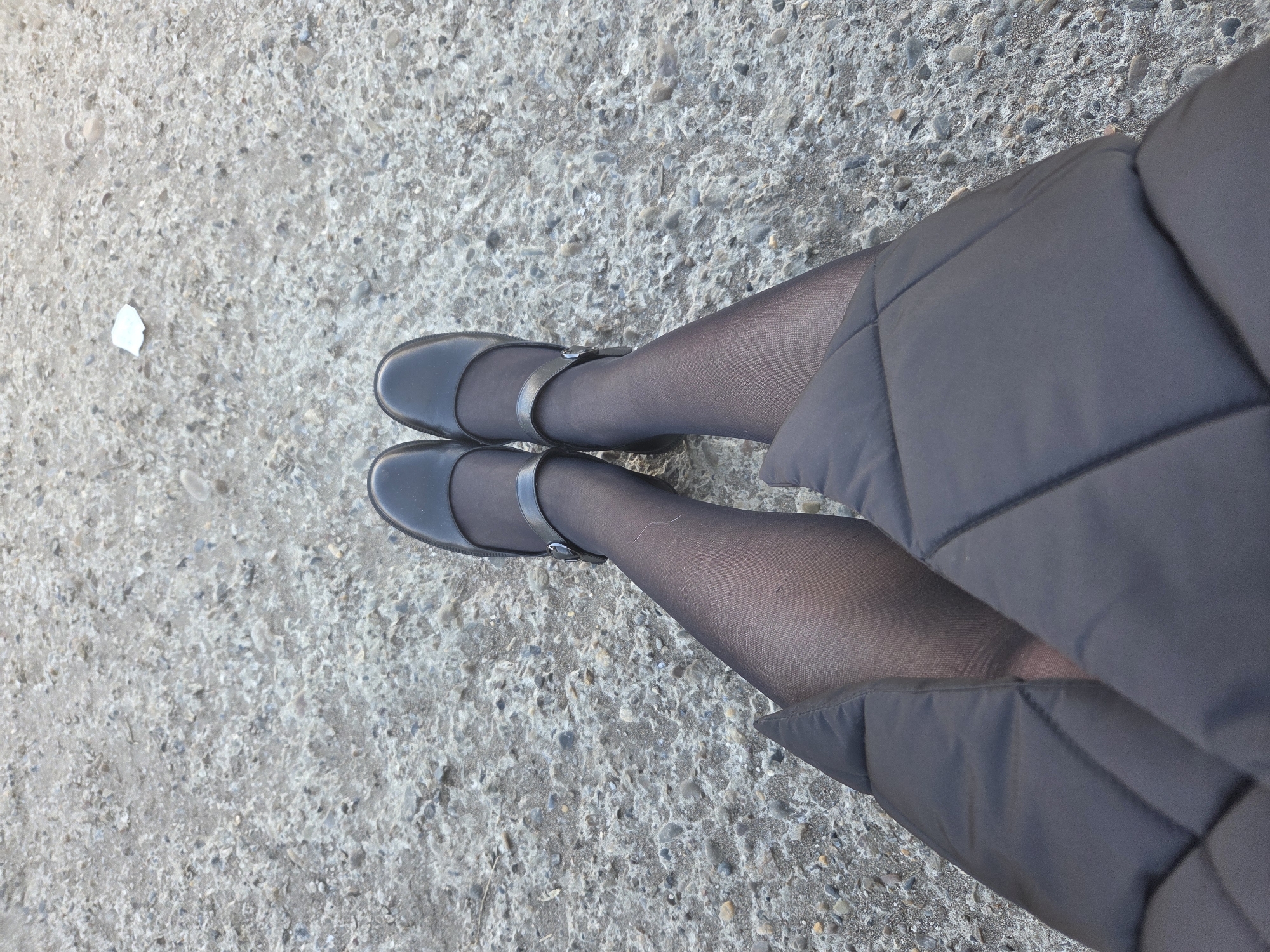 The image shows a downward-angled view of a person's legs and feet on a concrete surface. The person is wearing black tights and black Mary Jane-style shoes with a strap across the foot. A portion of what appears to be a padded, dark-colored coat or jacket is visible on the right side of the image, partially obscuring the legs. The background is a rough, speckled concrete surface. There is a piece of white trash in the upper left corner of the image. The city of Chaikovsky, Russia, is not discernible from the image.